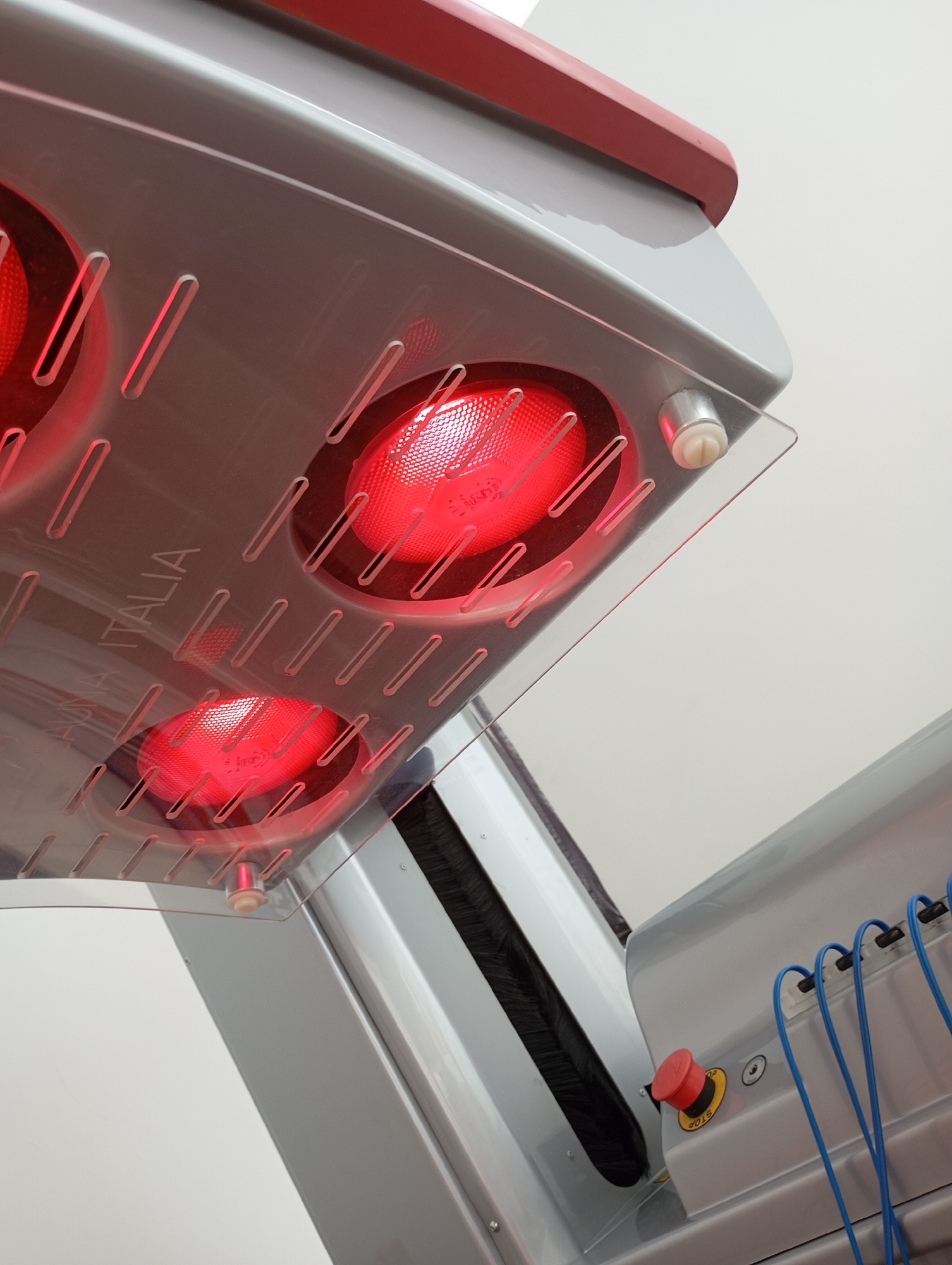 A close-up, low-angle interior shot of a tanning bed. The bed is silver with a red trim along the top edge. Inside, two red bulbs are visible through a clear plastic cover that has etched slots. The word "ITALIA" is etched into the plastic. A red emergency stop button is visible on the side of the bed. Blue wires are connected to the side of the bed. The background is a plain white wall. The image was taken in Novosibirsk, Russia.