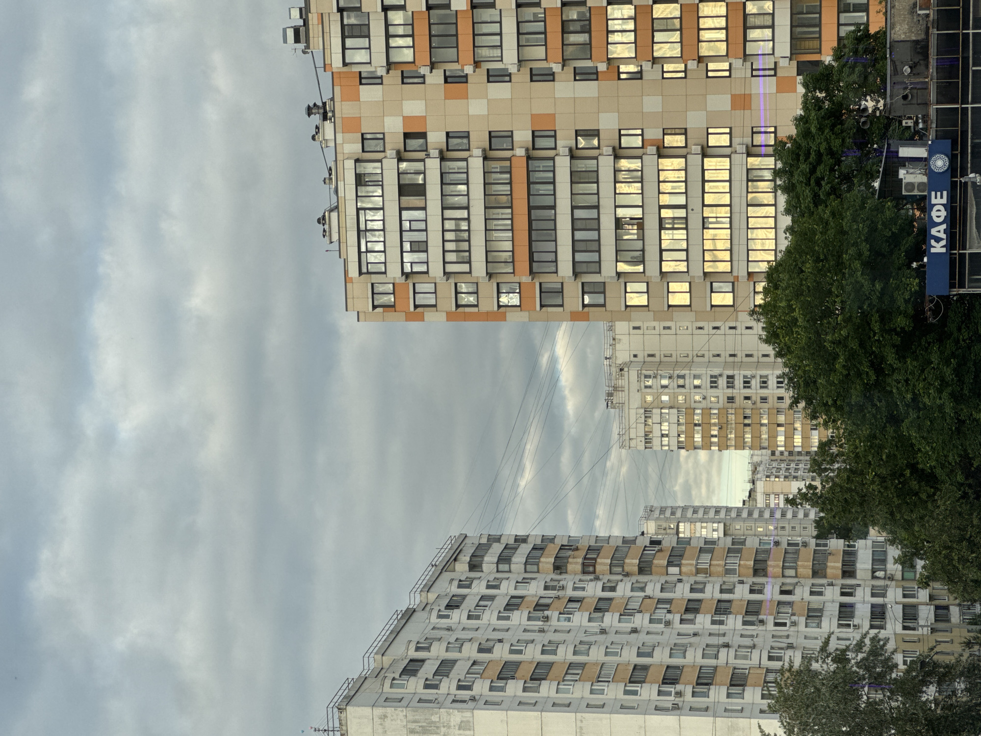 This image depicts an urban landscape in Moscow, Russia, dominated by several high-rise buildings under a partly cloudy sky. The scene is devoid of people.
In the foreground, on the left, an older, light grey high-rise building stands with numerous windows, some featuring air conditioning units, and intermittent light beige-brown panels below the windows. Its roofline is somewhat irregular. To the right, a more modern, tall building features a distinctive checkered facade of beige, orange, and white panels. Many of its windows exhibit a strong golden reflection, suggesting the time is either early morning or late afternoon, with sunlight breaking through the clouds.
Green foliage from trees partially obscures parts of the modern building on the right and also appears in the bottom left corner. A prominent blue sign with white Cyrillic text, reading "КАФЕ" (KAFE), meaning "cafe," is visible on the right, affixed to a structure near the modern building. A small circular logo is also present next to the text.
In the mid-ground and background, additional high-rise buildings are visible under the same overcast sky. Thin power lines crisscross the sky between the structures. The weather is partly cloudy, with moments of sunlight creating bright reflections. The overall setting is a bustling urban environment.