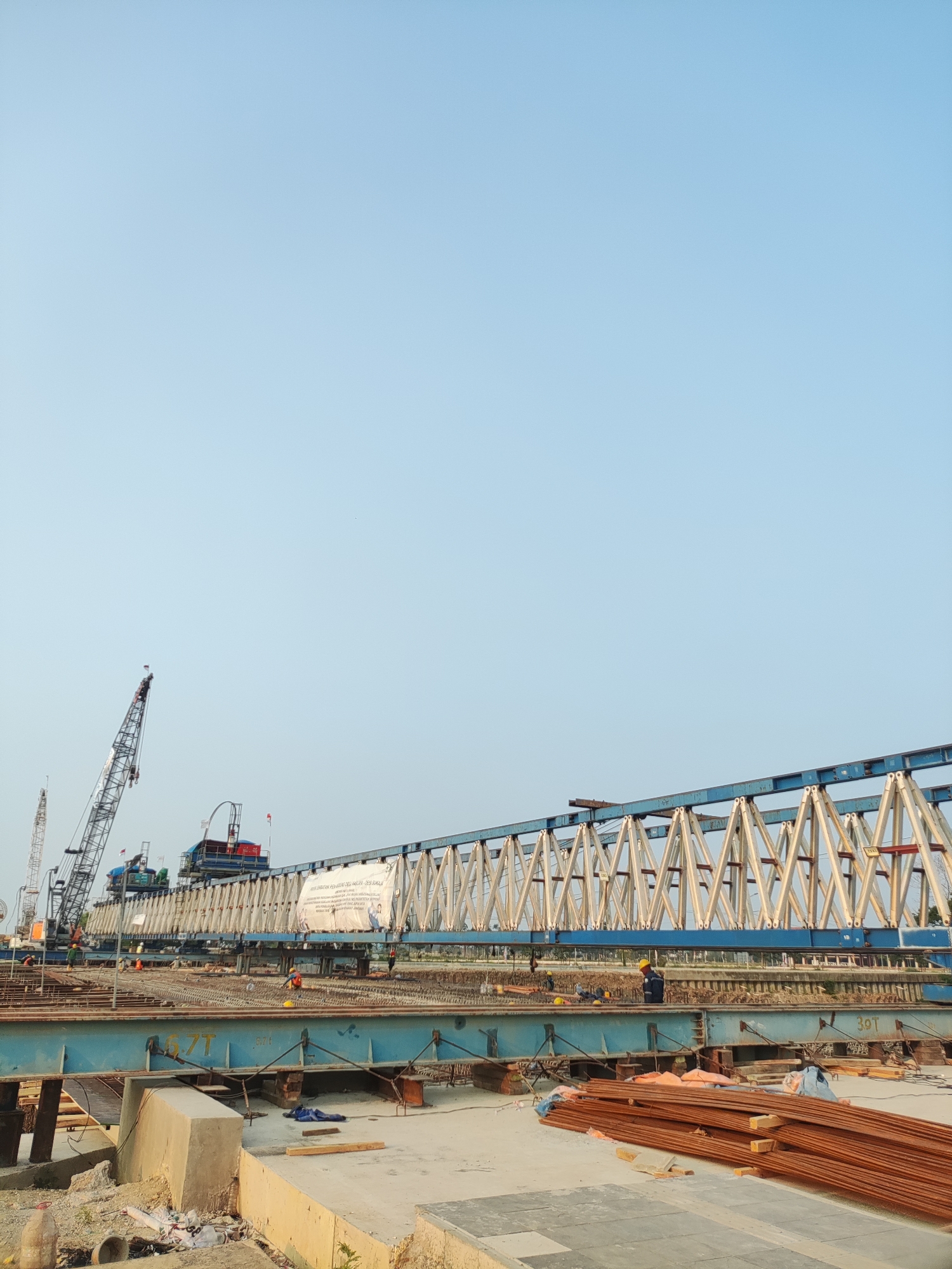 In Cikarang, Indonesia, a large-scale construction project is underway, likely for a bridge or elevated structure. A prominent feature is a long, blue steel truss spanning across the frame, supported by lighter-colored, triangular metal bracing. In the foreground, a concrete foundation and piles of rust-colored rebar are visible. Several construction workers, identifiable by their hard hats and safety vests, are present on the site, engaged in various tasks. A tall crane with a blue and white lattice boom is positioned to the left, suggesting heavy lifting operations are in progress. The sky is clear and a pale blue, indicating daytime. The overall scene depicts a dynamic and active construction environment.