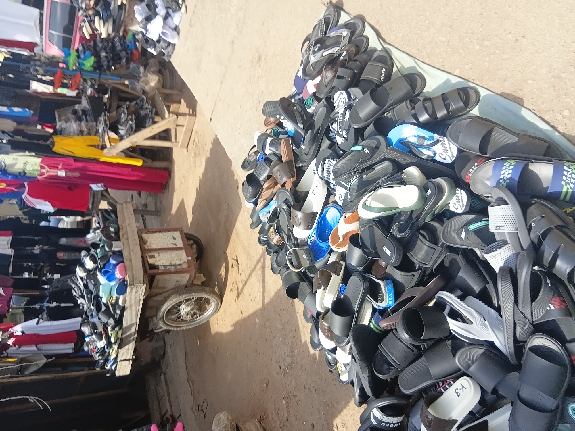 This is a forensic, matter-of-fact description of a street market scene in Jalingo, Nigeria. A large quantity of assorted footwear, predominantly sandals and flip-flops, is displayed on the ground, covered by a light blue tarp. The footwear is piled high, with various colors and brands visible, including black, white, brown, blue, and green options. Some pairs are neatly arranged, while others are in disarray.
Adjacent to the footwear display is a wooden cart, also laden with footwear. This cart has two wheels and appears to be a mobile vendor's stand. Behind the cart, a market stall is set up with clothing items hanging from racks. These clothes include t-shirts, dresses, and pants in a range of colors such as red, yellow, white, and black.
The ground is a sandy, dirt surface, indicative of an outdoor market. The lighting suggests a bright, sunny day. There are no visible people interacting directly with the footwear or clothing in the frame, though the context implies a commercial transaction is taking place. The environment is consistent with a bustling open-air market in an urban setting.