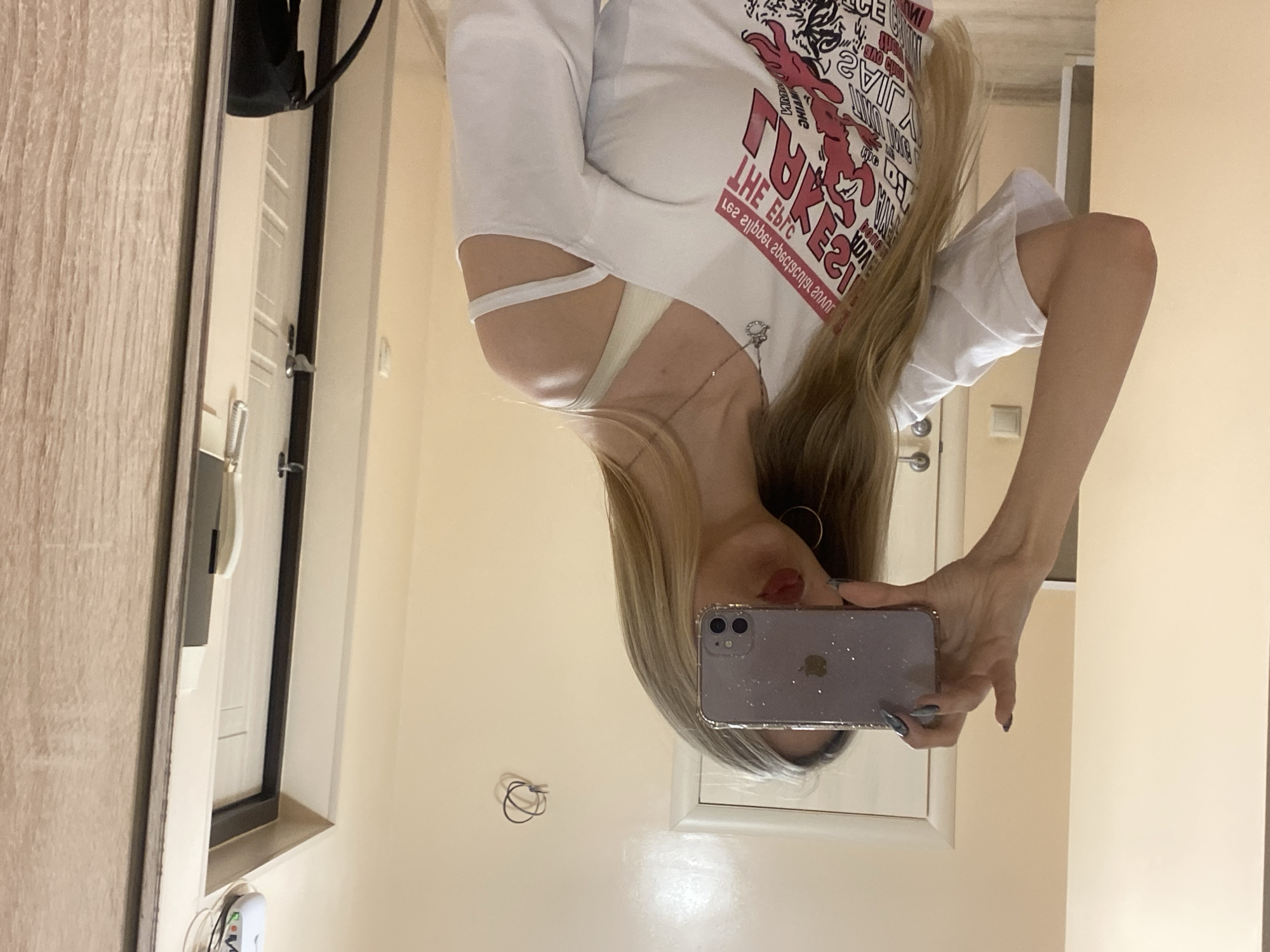 A woman is taking a selfie in a mirror. She is wearing a white long-sleeved shirt with a graphic design. She is holding a purple iPhone in her left hand. Her hair is long and blonde. She is wearing earrings. The mirror is located in a room with beige walls and a white door. There is a wooden door to the left. The location is Unknown, Unknown.