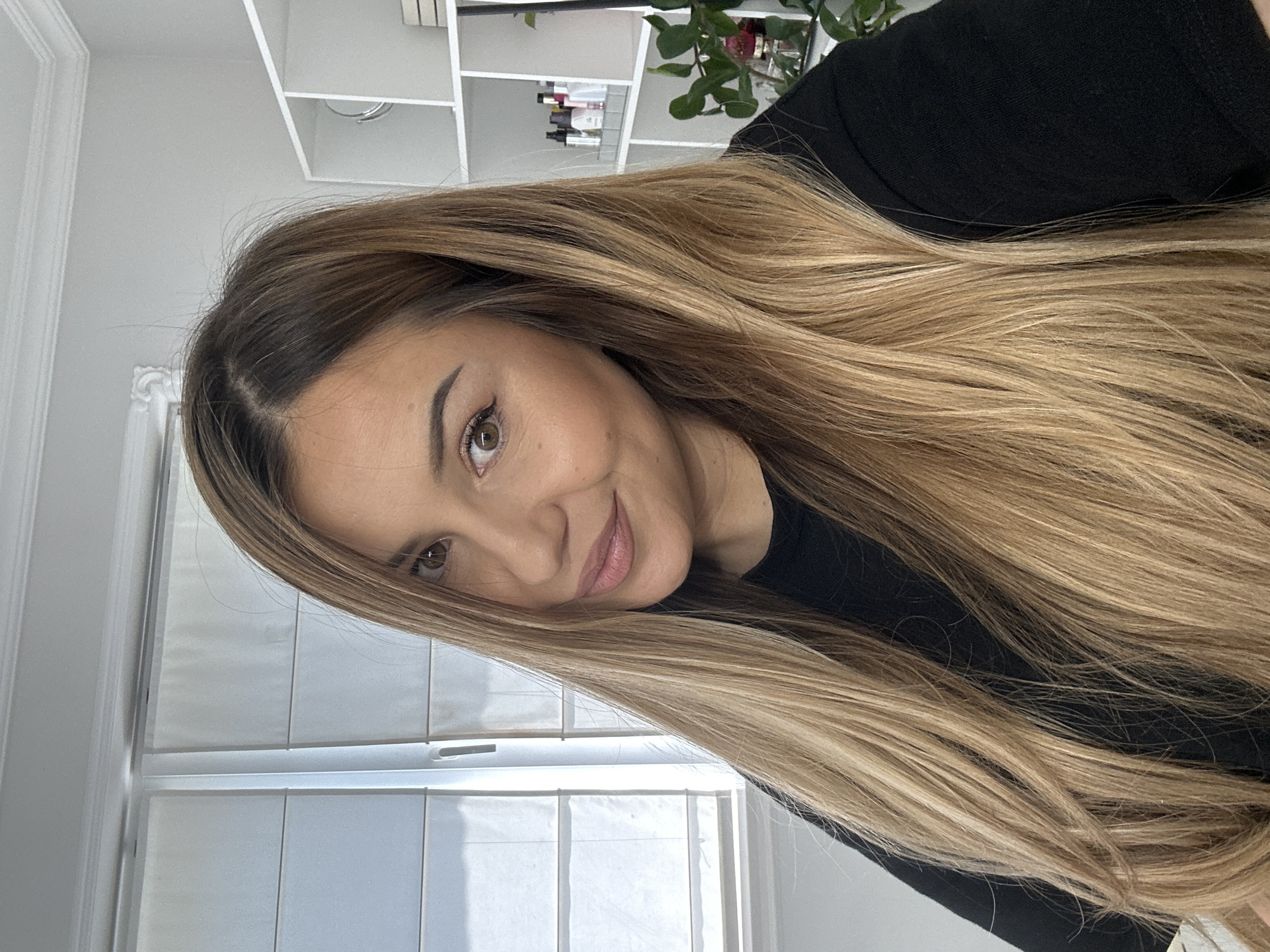 A medium close-up photograph depicts a young adult female, presumably in her 20s or 30s, inside a building in the city of Asenovgrad, Bulgaria. She has long, straight hair with a balayage color treatment, transitioning from medium brown at the roots to a golden blonde through the lengths and ends. Her hair is parted on the left side (from her perspective) and falls forward over both shoulders and chest. She has brown eyes, defined eyebrows, and appears to be wearing eyeliner and a soft pink lip color. Her expression is a gentle, slight smile, and her head is tilted subtly to her left. She is wearing a plain black long-sleeved garment. The background on the left side of the frame features a window fitted with white horizontal blinds, through which diffused natural light is entering the room. On the right side of the frame, a white shelving unit composed of multiple square compartments is visible. These shelves contain various small bottles and jars (likely cosmetics or toiletries), a circular mirror, and stacked items resembling books or decorative boxes. Below the shelving unit, a green leafy plant in a dark pot is positioned next to a reflective surface, possibly a decorative tray, which holds a small pink object. The overall lighting of the scene is bright and even.