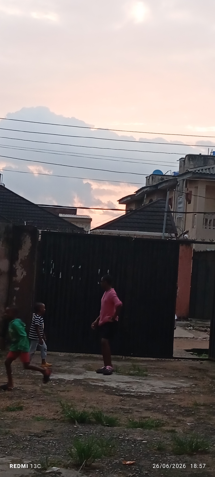The image depicts a group of children playing outside in what appears to be a residential area in Idimu, Nigeria. The scene is set against a backdrop of houses with dark tiled roofs and a tall black metal gate. Overhead, power lines are visible against a soft, hazy sky with hints of pink and orange, suggesting it is either sunrise or sunset.
Three children are the main subjects. On the left, a child in a green shirt and red shorts is running, their feet blurred with motion. To their right, another child, wearing a striped black and white shirt and light-colored pants, stands facing the gate. In the center-right, a taller figure, possibly an older child or young adult, is wearing a pink shirt and dark shorts and appears to be interacting with the younger children, perhaps playing or supervising. The ground is a mixture of dirt and gravel with patches of grass growing through.
The lighting and cloud colors suggest it's either dawn or dusk, casting a gentle, diffused light over the scene. The presence of residential buildings, the gate, and overhead wires are common features in urban or semi-urban Nigerian settings. The overall atmosphere seems relaxed and casual, indicative of children engaged in outdoor play. No specific event is clearly identifiable, but it's a snapshot of everyday life.
Visible text at the bottom left reads "REDMI 13C", indicating the phone model used to take the picture. At the bottom right, the date and time are displayed as "26/06/2026 18:57", which suggests the image was taken in the late evening.