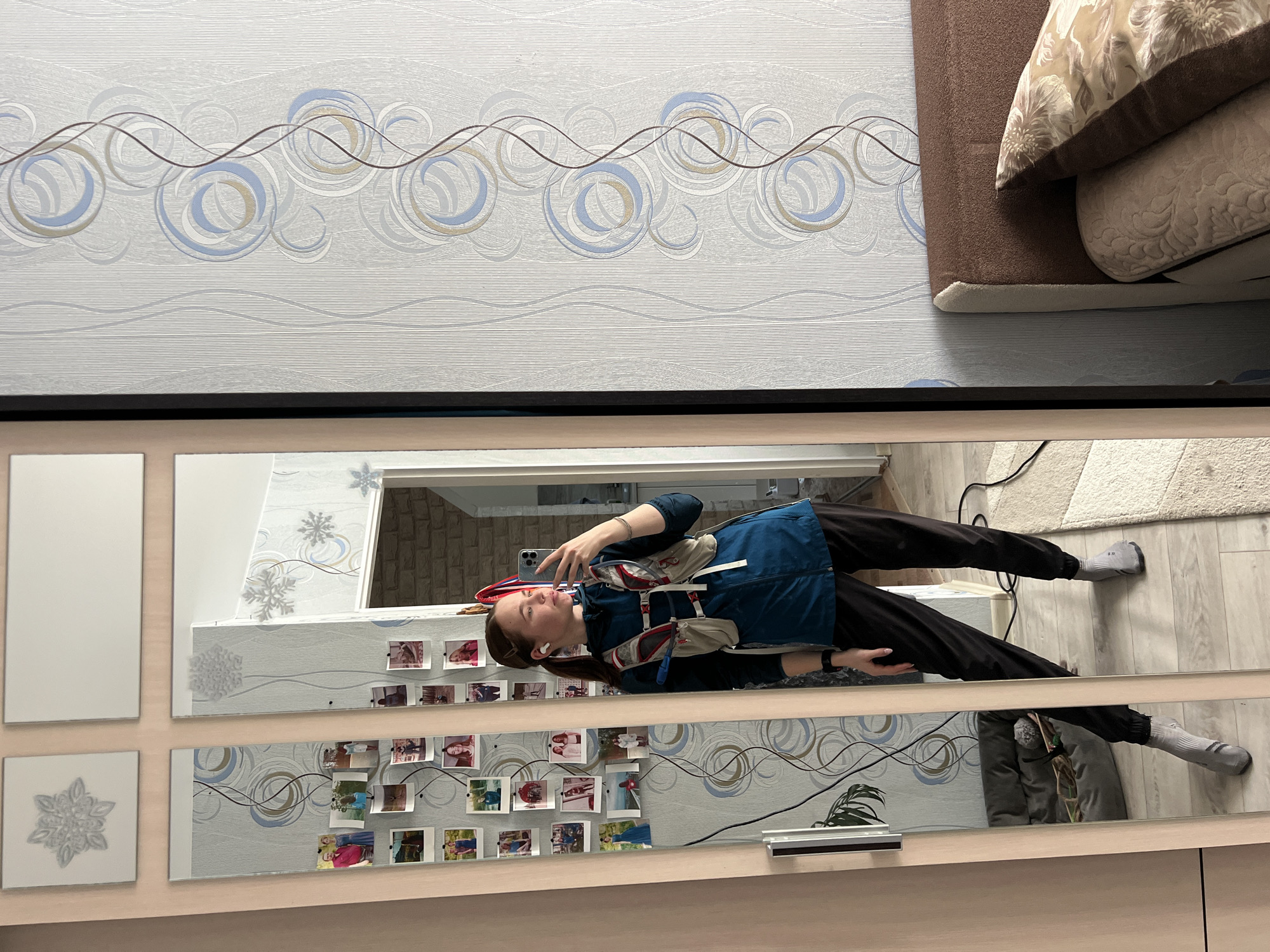 A woman stands in front of a mirror taking a selfie. She is wearing a blue jacket and black pants. Her hair is brown, and she has earbuds in her ears. The mirror reflects a room with wallpaper featuring a swirled pattern and photographs attached to the frame. A couch with patterned pillows sits at the top of the frame. The location is Chaikovsky, Russia.