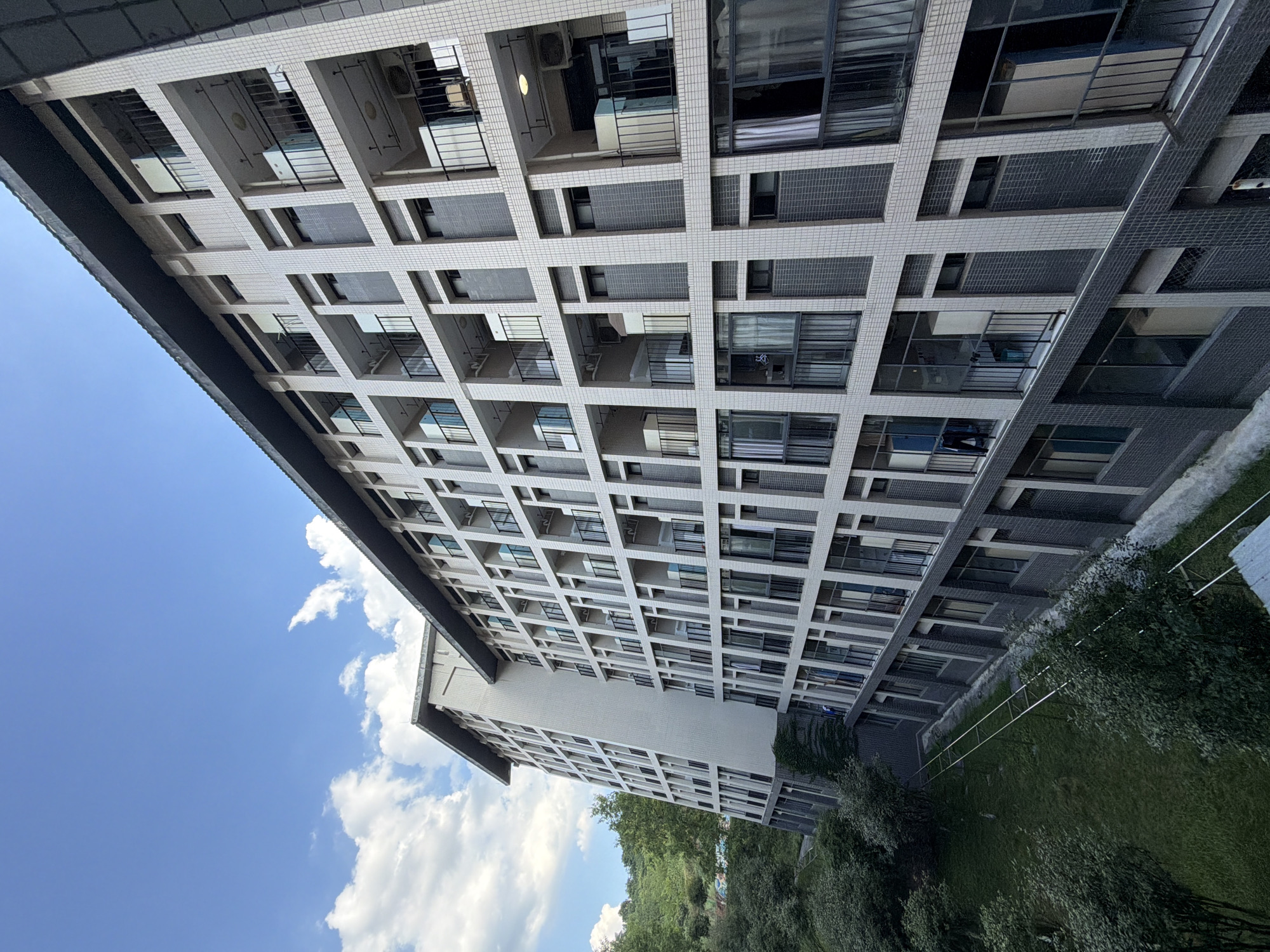 A drone shot captures a lengthy, multi-story building constructed with light-colored tiled facade and dark grey tiled accents, possibly a dormitory or apartment complex, situated in the city of Jiangjin, China. The building features numerous balconies with black metal railings and a repeating pattern of windows and recessed areas. Green trees and a grassy area flank the building on the left. The sky above is a clear blue with scattered white clouds. In the foreground, a narrow path or walkway with thin, horizontal metal poles is visible running alongside the grassy area. The building is angled, revealing its side facade.