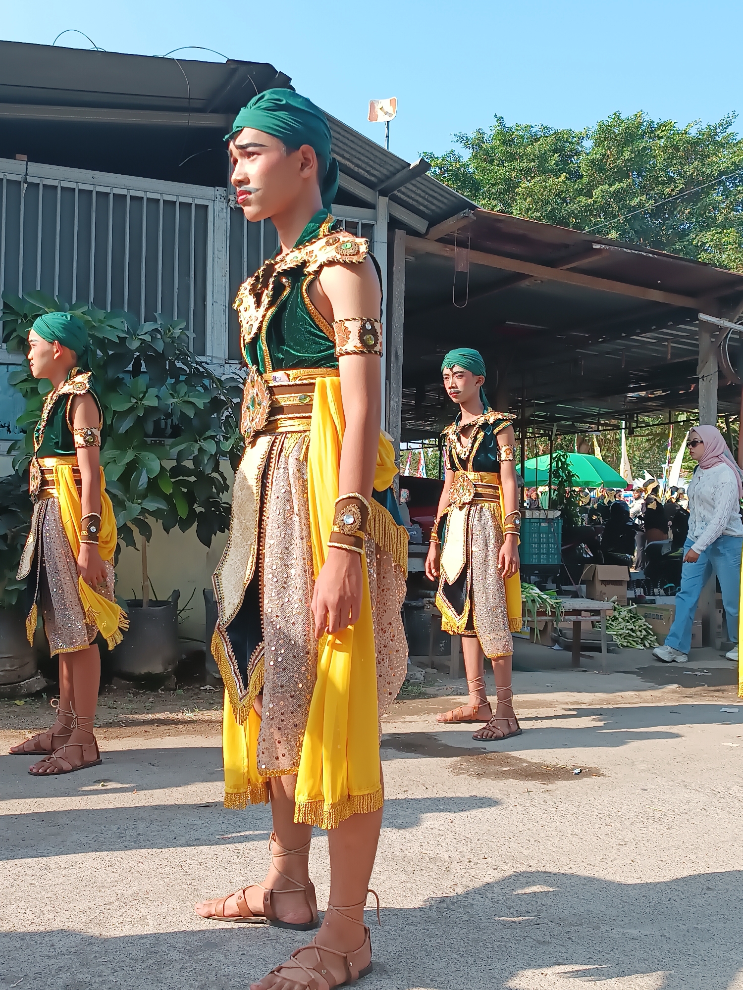 In Imogiri, Indonesia, three young males stand in a line, dressed in traditional Javanese attire. They wear green headscarves, elaborate gold and green vests with ornate collars, and knee-length skirts adorned with sequins and fringed yellow sashes. Their footwear consists of simple leather sandals with laces wrapped around their ankles. The central figure, facing left in profile, has makeup emphasizing his eyes and lips, along with a painted mustache.
They are positioned outdoors on a paved surface, likely a street or pathway, under a bright blue sky. Behind them, to the left, is a building with a corrugated metal roof and barred windows, partially obscured by lush green foliage in large planters. To the right, a covered market area is visible, with stalls, tables, and shoppers. A woman in a light blue shirt and jeans, wearing a pink headscarf, walks away from the camera in the background. The overall scene suggests a cultural event or performance taking place in a semi-urban or market setting.