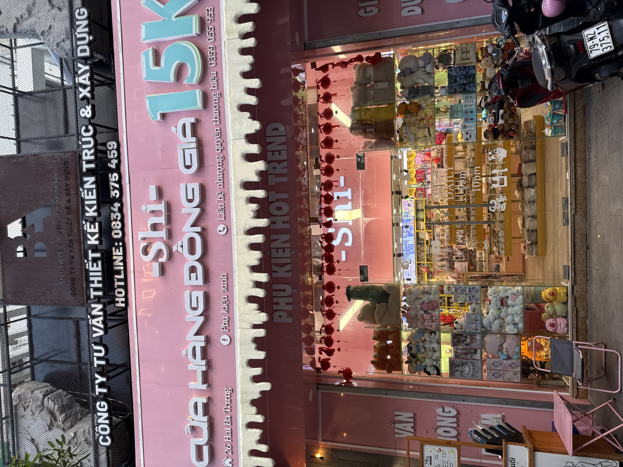 A storefront in Nha Trang, Vietnam, is captured in this medium-shot, eye-level photograph. The store's facade is painted a bright pink and features prominent signage. The upper portion of the building displays text in Vietnamese, including "CÔNG TY TƯ VẤN THIẾT KẾ KIẾN TRÚC & XÂY DỰNG" and a hotline number. Below this, a large pink sign reads "CỬA HÀNG ĐỒNG GIÁ" with the large turquoise characters "15K" to the right. An address, "26 Hai Bà Trưng," is visible on the left, and a Facebook page name, "Phụ kiện xinh," is on the right, indicating the store sells affordable items.
The storefront is glass-enclosed, revealing a display of plush toys and merchandise. The interior appears to be well-lit and organized with shelves stocked with various items, including stuffed animals, dolls, and gift items. A sign inside indicates the store's operating hours: "DAILY 9am - 10pm". Red lanterns are suspended above the entrance, adding a decorative touch.
In front of the store, two motorcycles are parked on the pavement. One is a dark red scooter, and the other is a black scooter with a visible license plate reading "79-N2 375.11". A small pink table and a grey folding chair are placed to the left of the entrance. The pavement in front of the store is made of grey concrete slabs. The overall impression is of a busy commercial street scene in Nha Trang.
