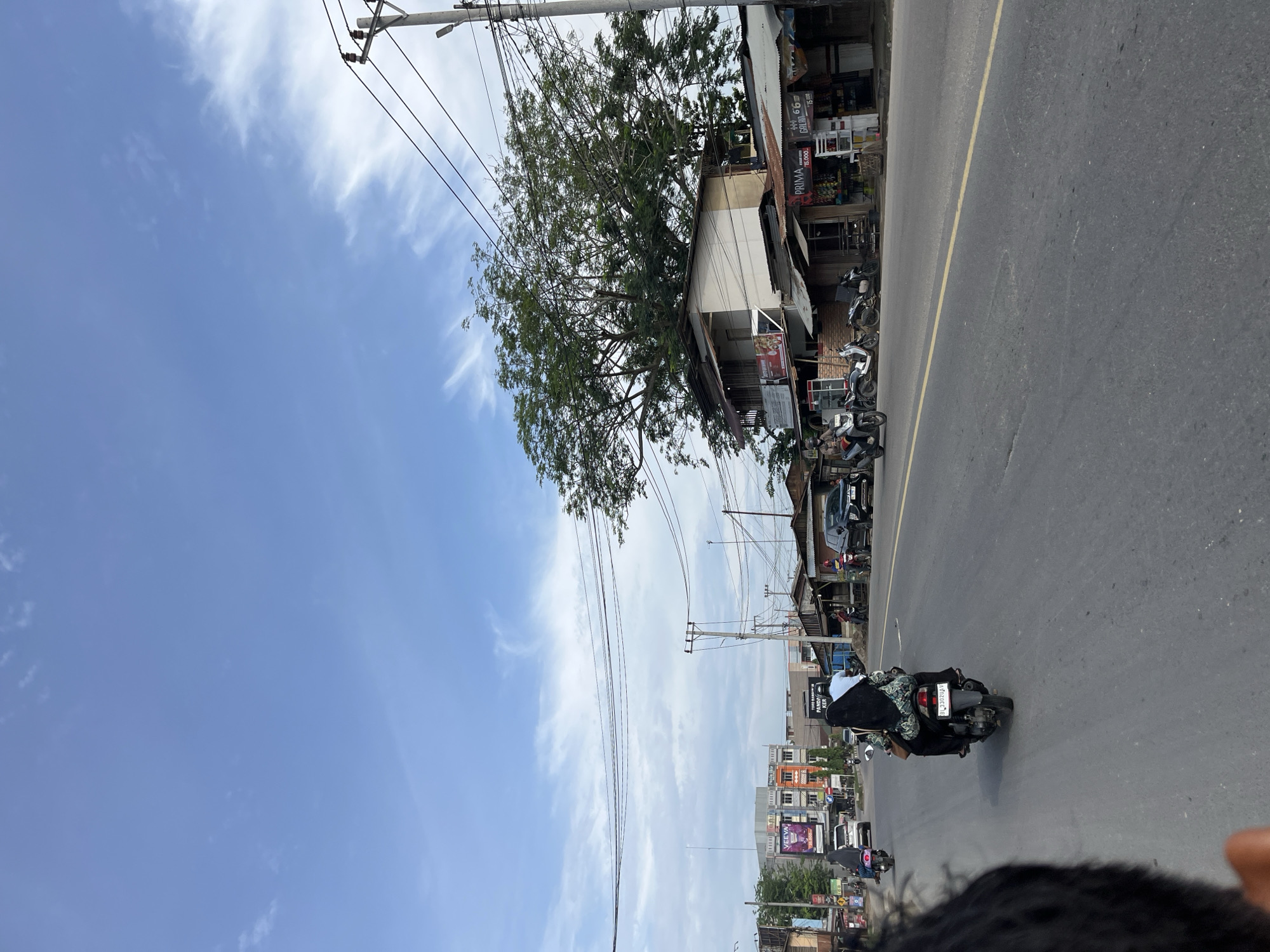 The image depicts a street scene in Sukajadi, Indonesia, under a cloudy blue sky. A paved road with a yellow dividing line dominates the foreground. Several motorcycles are visible, with two in the immediate foreground. One motorcycle carries two individuals, the passenger wearing a headscarf and patterned jacket, and the rider in a dark outfit. Another motorcycle to the left has a single rider in a brown jacket. Further down the road, other motorcycles and a black SUV are in motion.
The roadside is lined with buildings and shops. On the left, a multi-story building with colorful signage, including a large "VEEVA" advertisement, stands out. Adjacent to it are smaller, more traditional structures with tin roofs. To the right, a prominent two-story building with a large tree overshadowing it is visible. Various shops, some with awnings and displays of goods, are situated along this side of the street, with signs such as "PRIMA" and "GALON 66" visible. Utility poles with a network of electrical wires are present throughout the scene. The overall impression is that of a bustling, urban commercial area.