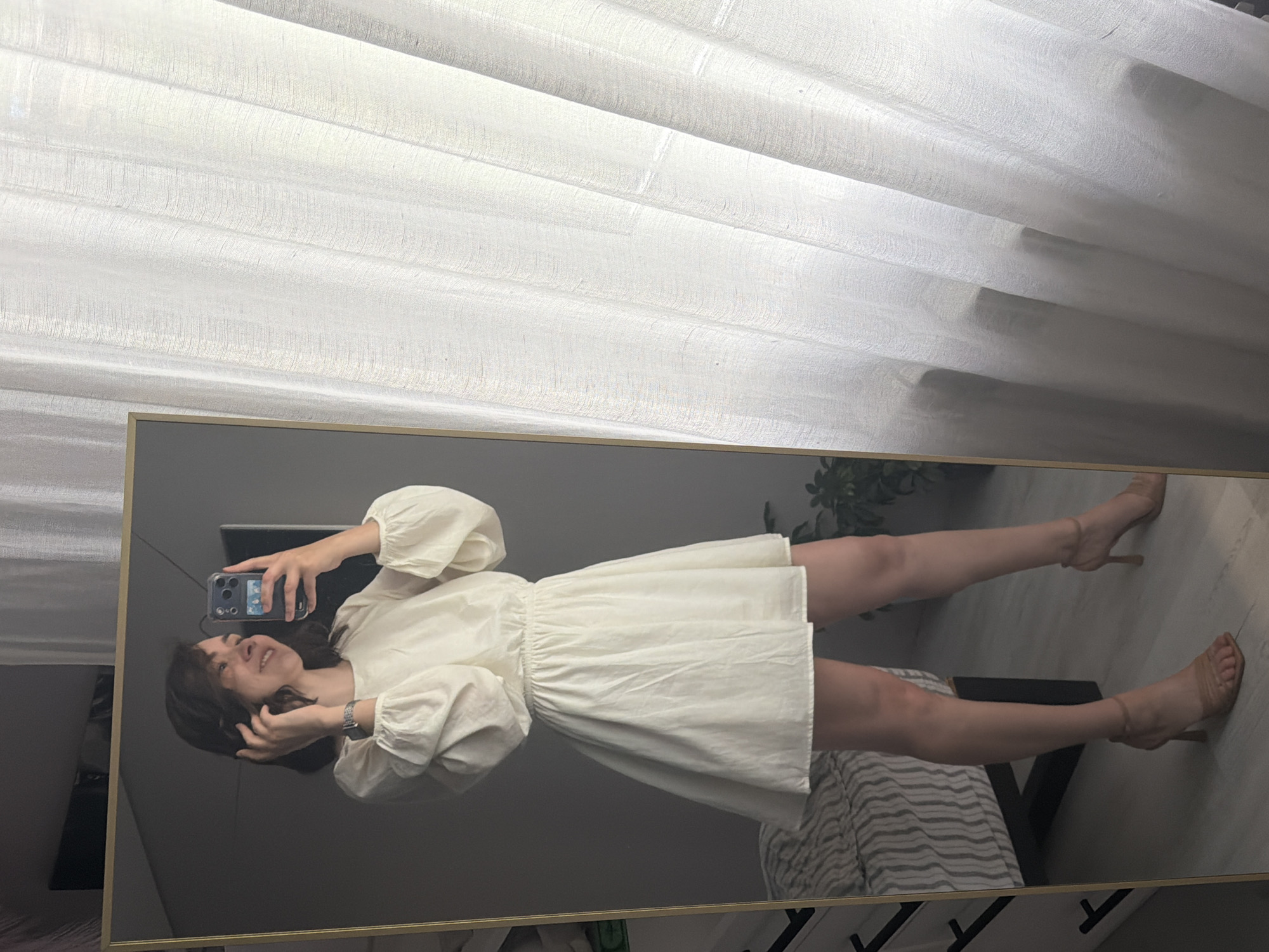In a well-lit indoor setting, likely a bedroom in Chaikovsky, Russia, a young woman stands facing a large, gold-framed mirror, taking a selfie with her phone. She is wearing a cream-colored, short-sleeved dress with voluminous sleeves and an elasticated waist, paired with nude-colored heeled sandals. Her hair is short and brown, and she is smiling as she touches her hair. The reflection in the mirror shows the woman from the waist up, as well as her legs and feet. In the background of the mirror's reflection, a bed with striped bedding is visible, along with a plant. To the right of the woman, a sheer white curtain is drawn, filtering natural light into the room. The overall impression is one of casual elegance and a moment of self-appreciation.