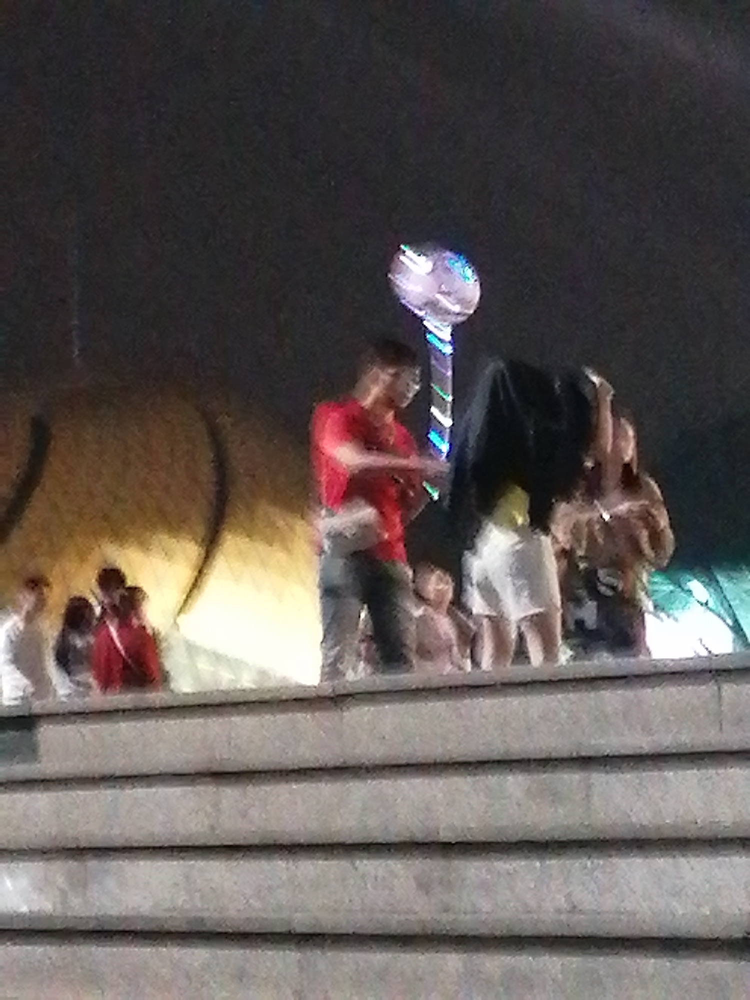 In this nighttime outdoor scene, a group of people are gathered on a set of concrete steps. The background features a blurry, illuminated structure with a large, curved, golden-yellow surface, possibly a building or art installation. Above the crowd, a colorful, glowing orb on a pole is visible, suggesting a festive atmosphere, perhaps a fair or celebration.
The primary subjects are several individuals. On the left, a person in a white shirt stands. To their right, a group of people, some in red shirts, are clustered together. In the center of the image, a man in a red shirt and blue jeans is prominent, interacting with something covered by a dark cloth, held by another person. To the far right, a woman in a patterned top is also visible. There are hints of younger individuals among the group, possibly children.
The activity appears to be a performance or presentation, given the presence of the covered object and the onlookers. The overall mood is one of public gathering and entertainment. The lighting suggests it is night. While the specific location in Da Lat, Vietnam, is provided, no distinct geographical landmarks are clearly identifiable within the image itself. There is no visible text in the image. The focus is on the human interaction and the ambient light of the event.