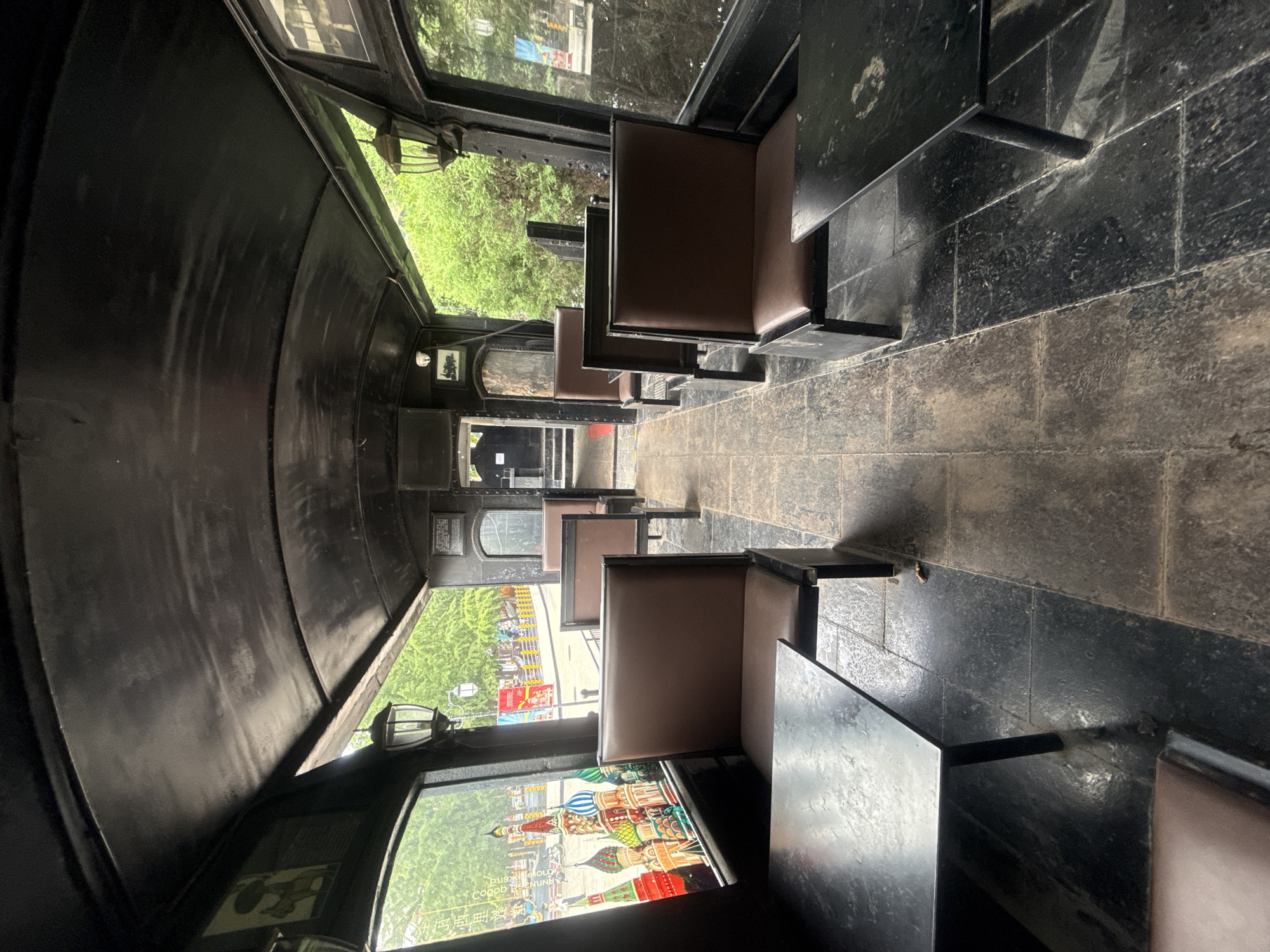 The photograph shows the interior of a refurbished train carriage, seemingly repurposed as a dining establishment, in Qinhuangdao, China. The carriage is dimly lit, with dark, black walls and a curved, black ceiling. Rows of brown, upholstered seats with metal frames flank a central aisle. Small, dark-colored tables are positioned in front of some of the seats.
Large windows run along the sides of the carriage, offering views of the outdoors. Through the windows, trees and greenery are visible, along with elements of an urban environment, including streetlights and distant buildings with colorful domes, reminiscent of St. Basil's Cathedral in Moscow, suggesting a themed attraction. The flooring appears to be made of dark stone tiles, showing signs of wear. Decorative lamps, in a vintage style, are mounted on the walls near the windows. The overall impression is of a quirky, themed seating area.