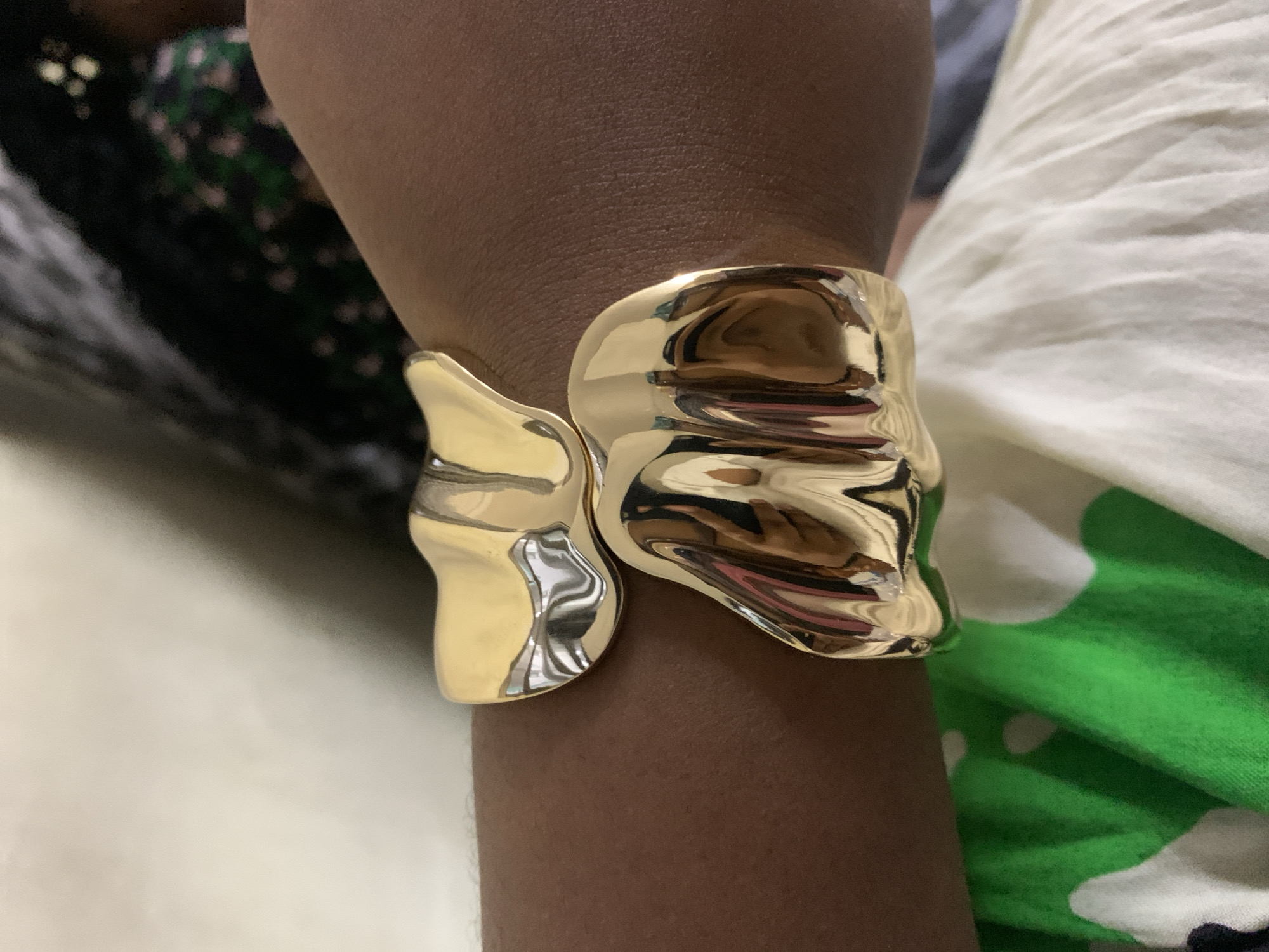 A close-up shows a person's wrist adorned with a gold-colored bracelet. The bracelet has an irregular, abstract shape with a shiny, reflective surface. The person's skin tone is medium-dark. Part of a garment with a black and green floral pattern is visible on the left. On the right, there's another garment with a white base and green patterns. The background is blurred and indistinct. The image does not provide enough information to determine if the location is Dar es-Salaam, Tanzania.