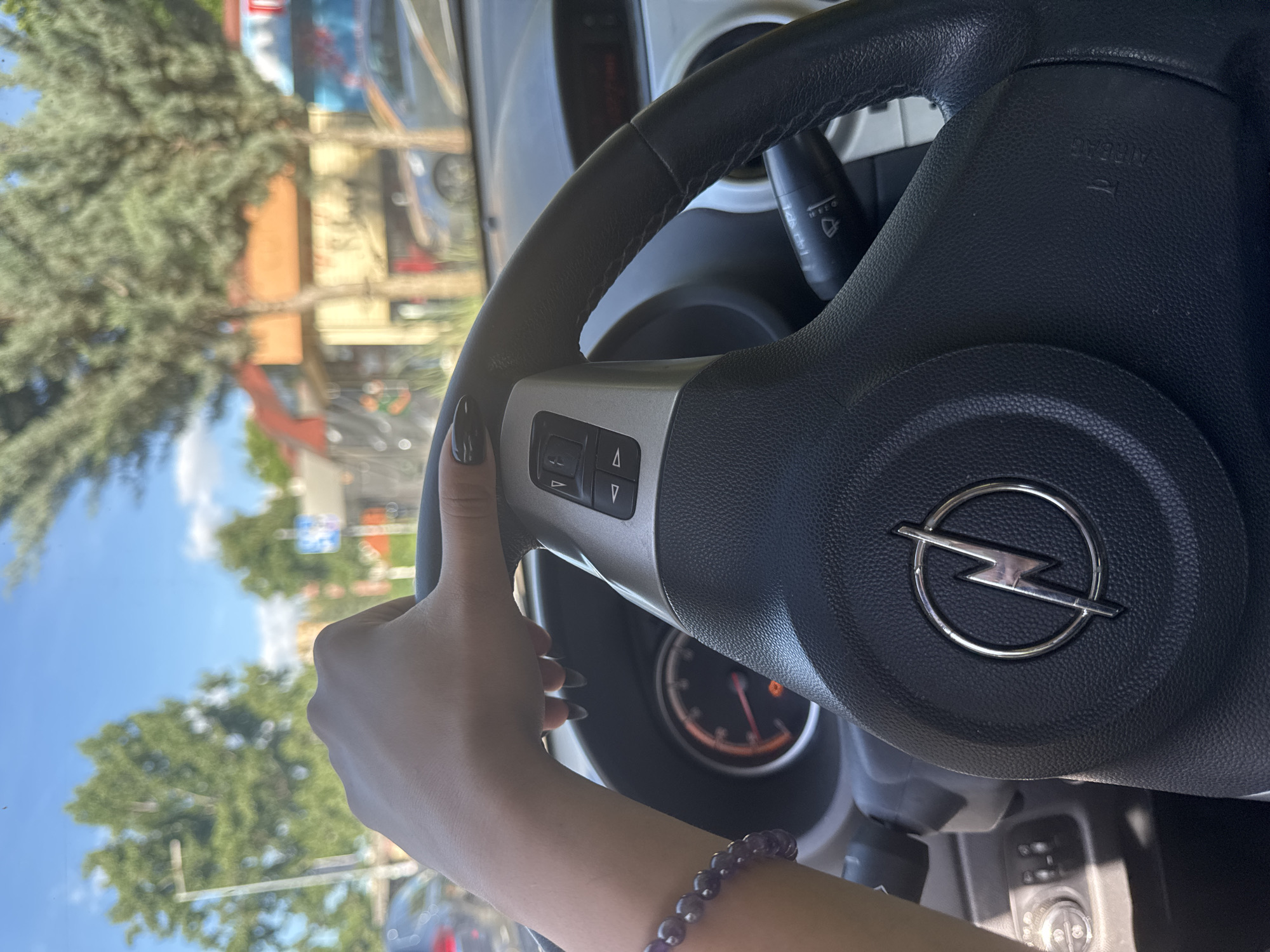 A partial view of the interior of a car is visible, focusing on the steering wheel and a person's left hand gripping it. The steering wheel features the logo of Opel. The hand has long, pointed nails painted black. A purple beaded bracelet adorns the wrist. The dashboard and part of the windshield are visible, with a blurry view of what appears to be a city street through the windshield. The vegetation and architecture suggest a location in Sofia, Bulgaria. Buildings are partially visible, along with a street with cars parked. The perspective is from within the vehicle, suggesting the photo was taken by someone inside.