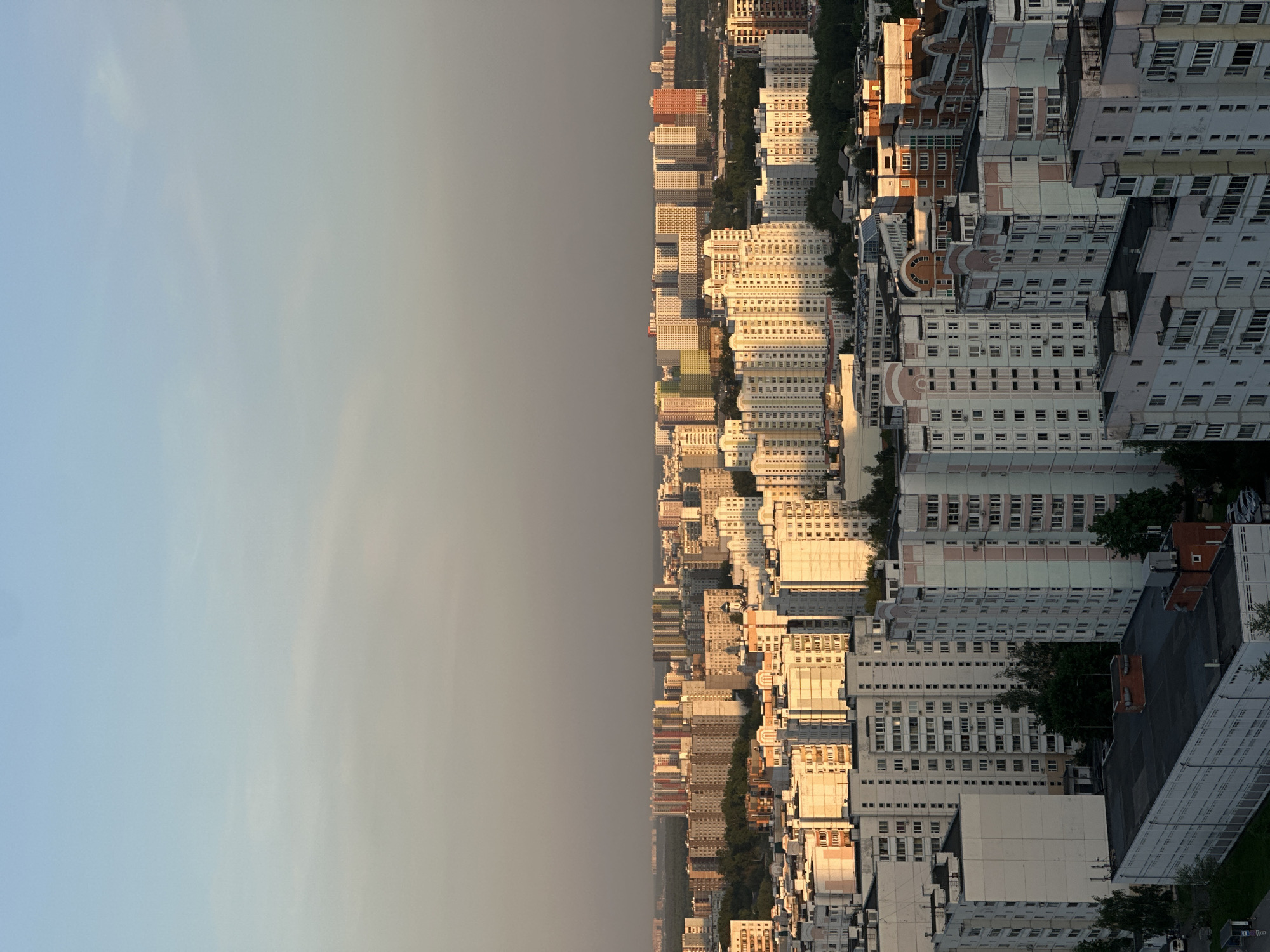 This image shows a cityscape under a hazy sky. The architecture consists primarily of residential buildings, possibly apartment complexes, characterized by their uniform appearance and numerous windows. The buildings are situated closely together, suggesting a dense urban environment. The lighting indicates either early morning or late afternoon, with some buildings catching the golden sunlight. The overall impression is of a bustling urban area.
