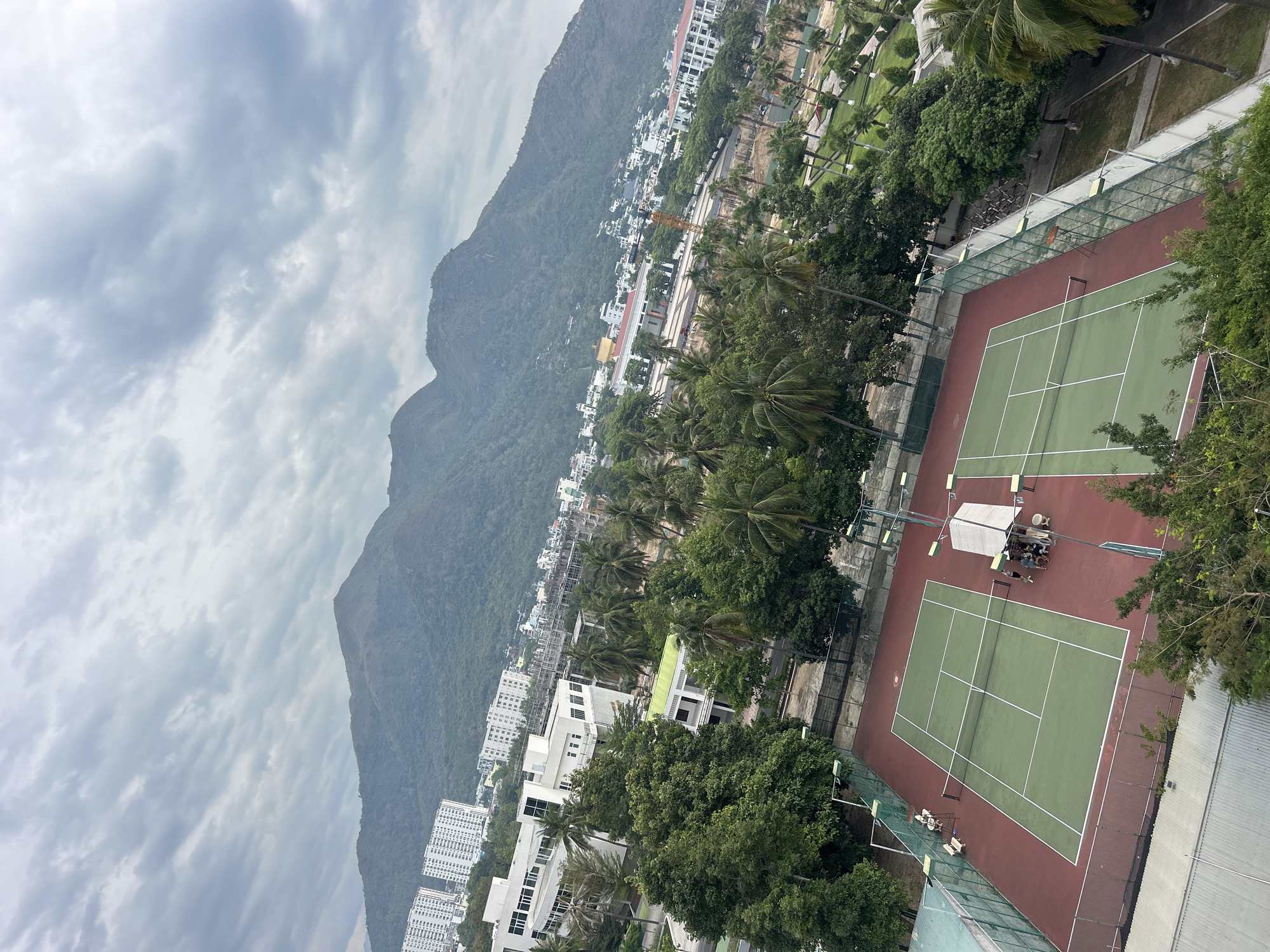 This high-angle, landscape-oriented photograph shows tennis courts with mountains and buildings in the background in Bac Nha Trang Ward, Vietnam. The sky is overcast with gray and white clouds. A large mountain is located on the left, extending partially into the city. The city contains a mix of low-rise and high-rise buildings, with visible construction. Palm trees and other greenery are scattered throughout the cityscape. To the right are two tennis courts, surrounded by green mesh fencing. Several individuals are visible on one of the courts, appearing to be taking a break under a shaded structure with tables and chairs.