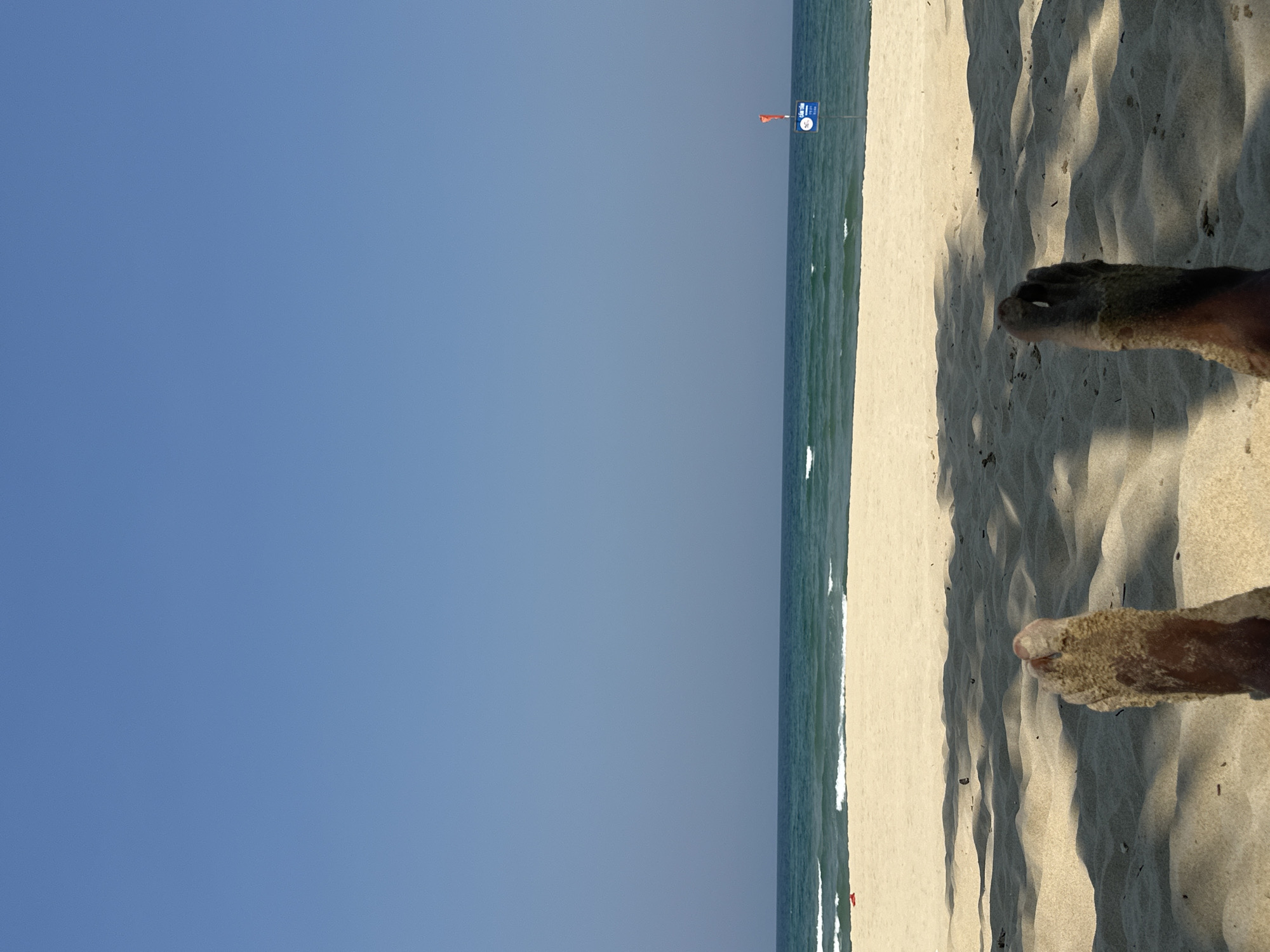 A seaside scene in Đà Nẵng, Vietnam features a blue sky, an ocean with small waves, and a sandy beach. A warning sign with an orange flag is visible along the shoreline. Two feet are partially buried in the sand in the foreground. Tree shadows fall across the sand.