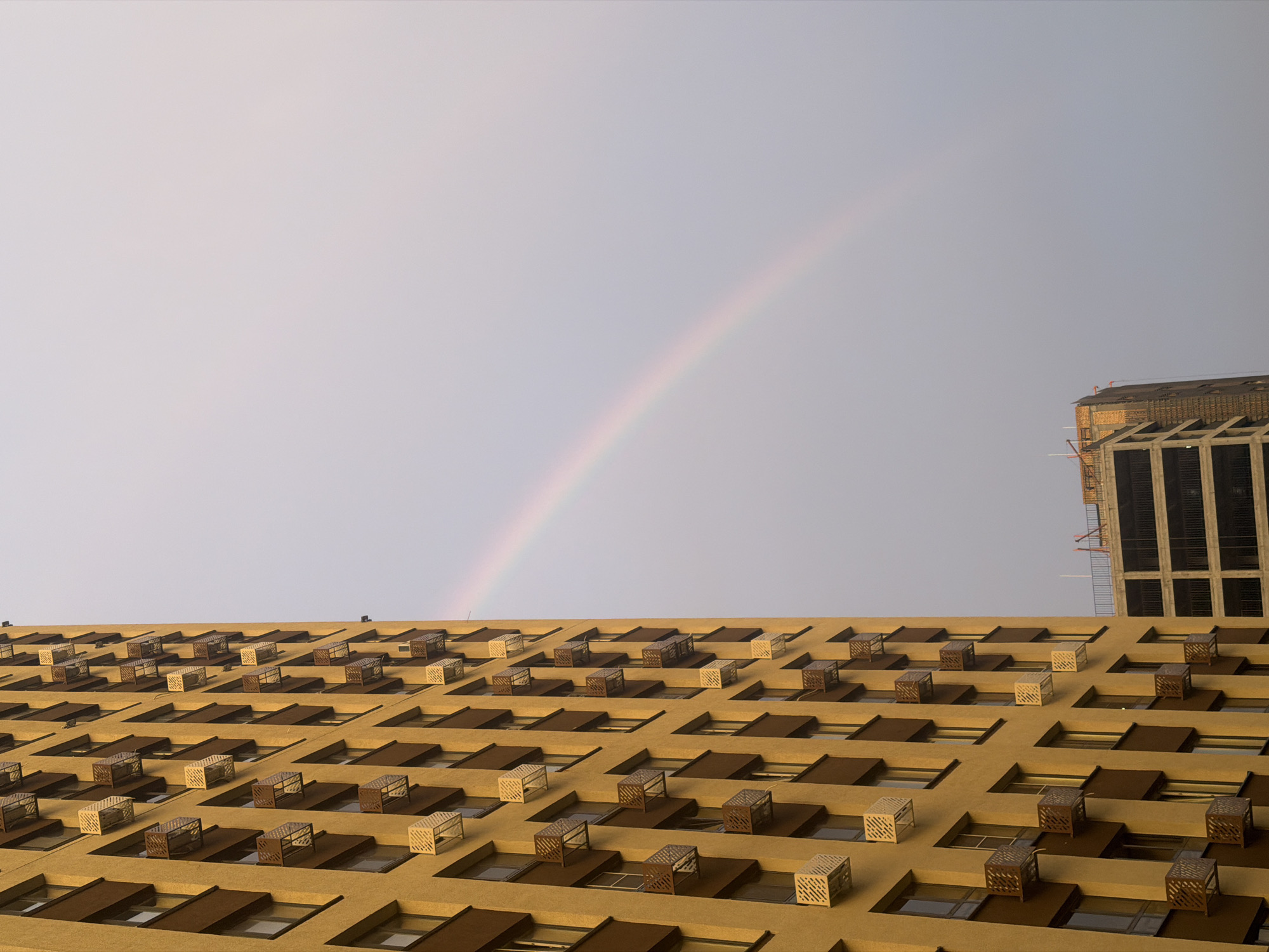 In Kazan, Russia, a golden-hued residential building stands tall, its facade adorned with numerous identical window units or external air conditioning enclosures, arranged in a grid pattern. To the right of this building, a fainter, partially visible rainbow arcs across a pale, overcast sky. In the background, another building, appearing to be under construction, reveals its concrete framework and scaffolding. The scene suggests an urban environment, possibly after a rain shower, with the natural phenomenon of a rainbow juxtaposed against modern architecture and ongoing development.
