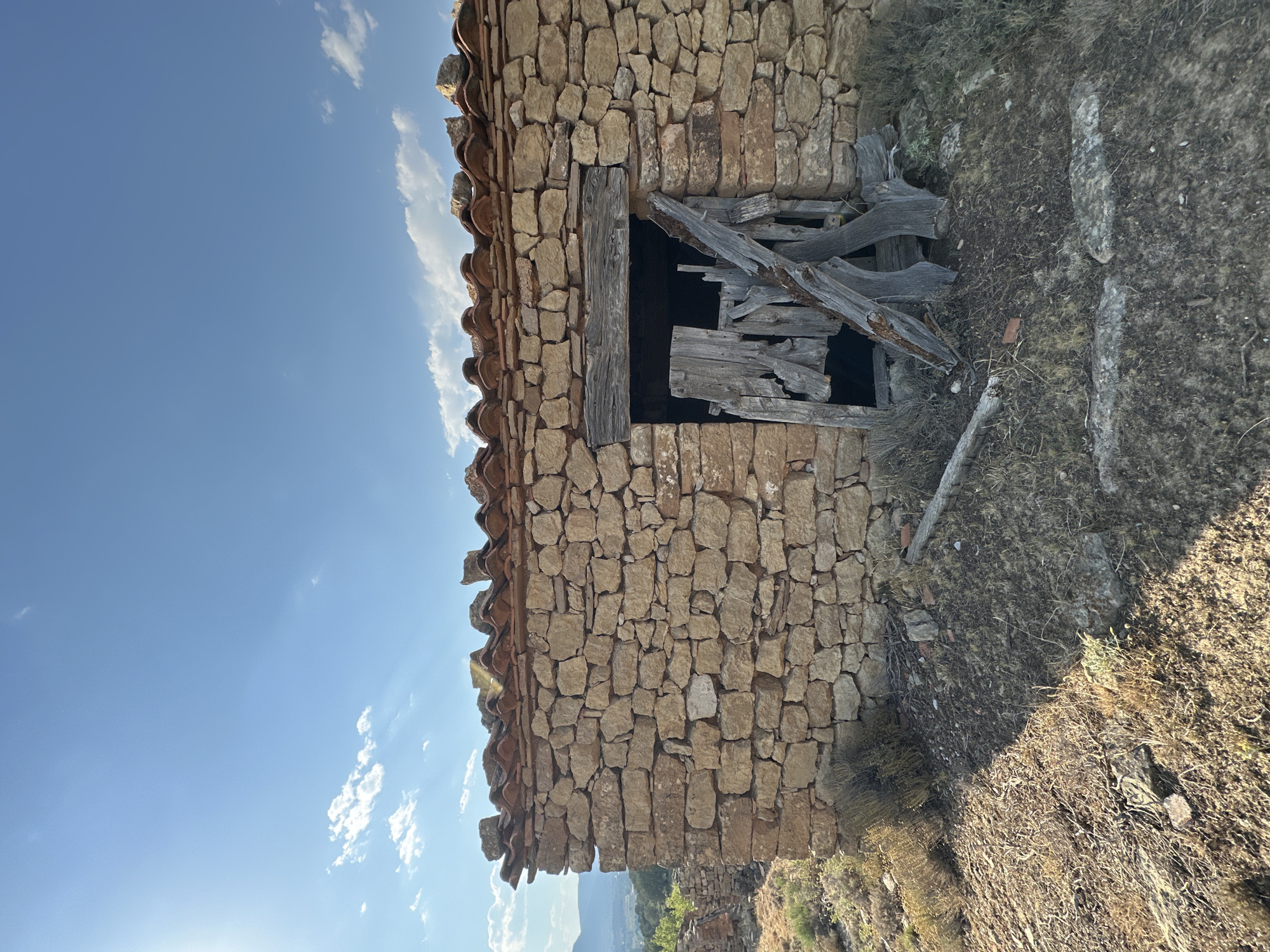 The image depicts a small, dilapidated stone structure with a terracotta tile roof situated on a hillside. The structure has a large, open doorway filled with broken wooden debris, suggesting disrepair. The surrounding environment is dry and arid, with sparse vegetation and loose stones. In the background, a hazy landscape of hills and distant mountains is visible under a clear blue sky with scattered clouds. The sun is shining brightly, casting shadows across the ground. The style of the stonework and roofing is consistent with traditional rural architecture found in the region of Alpuente, Spain.