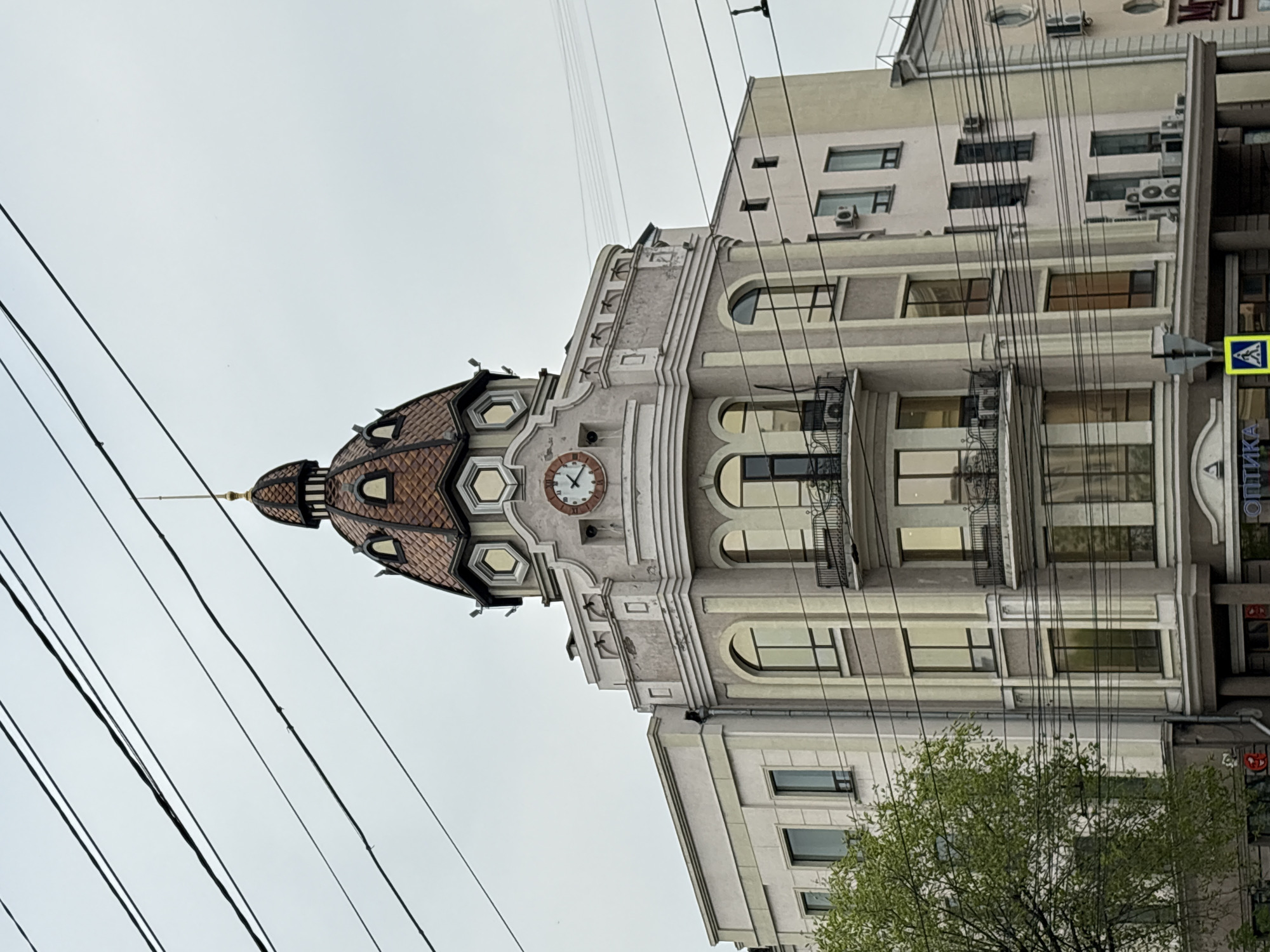 The image shows a building with a distinctive domed tower, likely located in Khabarovsk, Russia, based on the architectural style and the Cyrillic script visible on a sign. The building has multiple floors, with light-colored facades and a clock on the tower's face. Electrical wires are prominent in the foreground, crisscrossing the sky. A tree with green foliage is visible at the bottom right of the frame. The sky appears overcast.