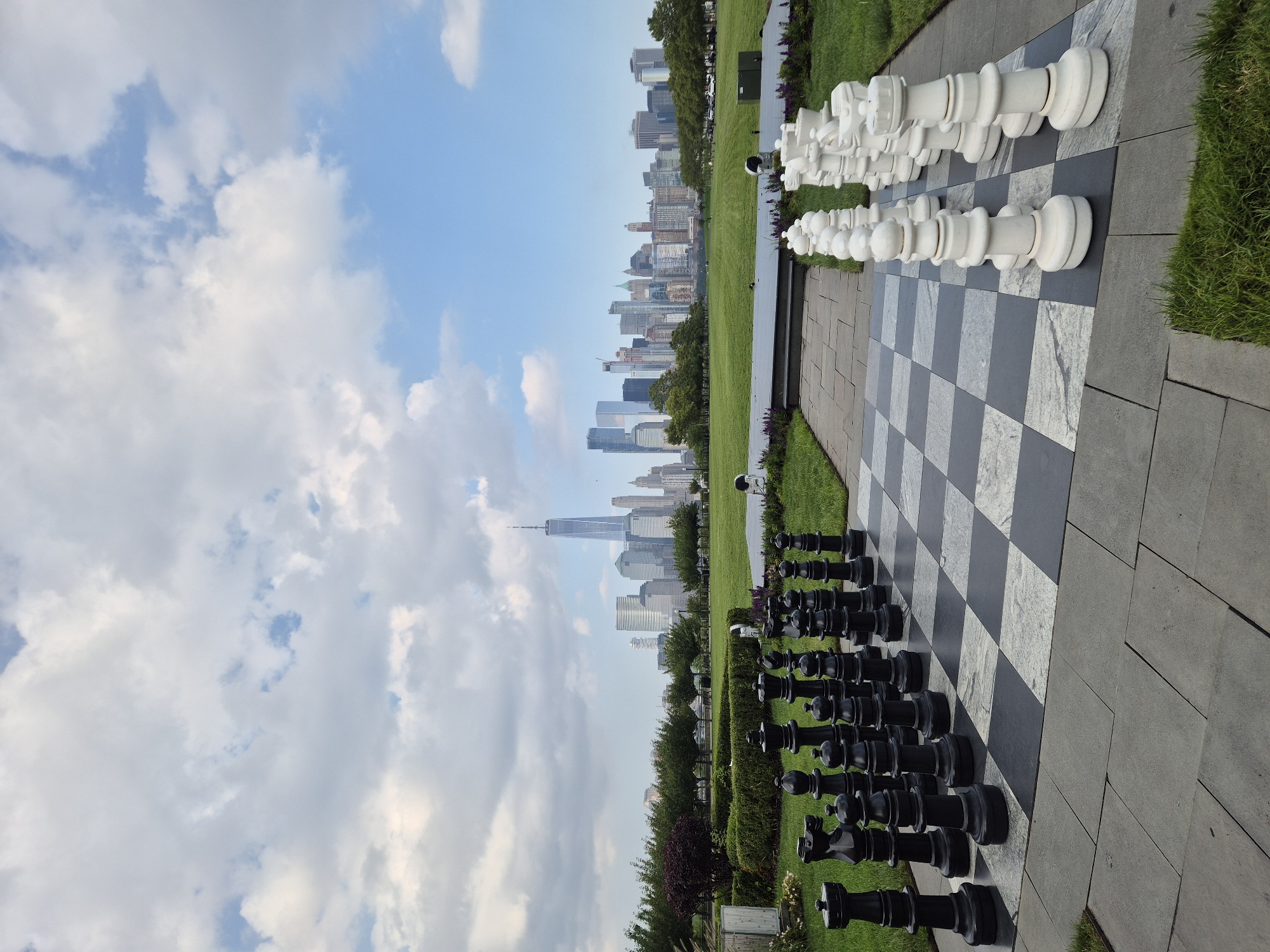 The image depicts a large outdoor chessboard with oversized black and white chess pieces set up in Liberty State Park in Jersey City, New Jersey. The iconic New York City skyline, including the One World Trade Center, serves as the backdrop. The sky is partly cloudy with patches of blue visible. The chessboard is situated on a paved area surrounded by manicured green lawns and some shrubbery. There are no people visible in the scene. The lighting suggests it is daytime.