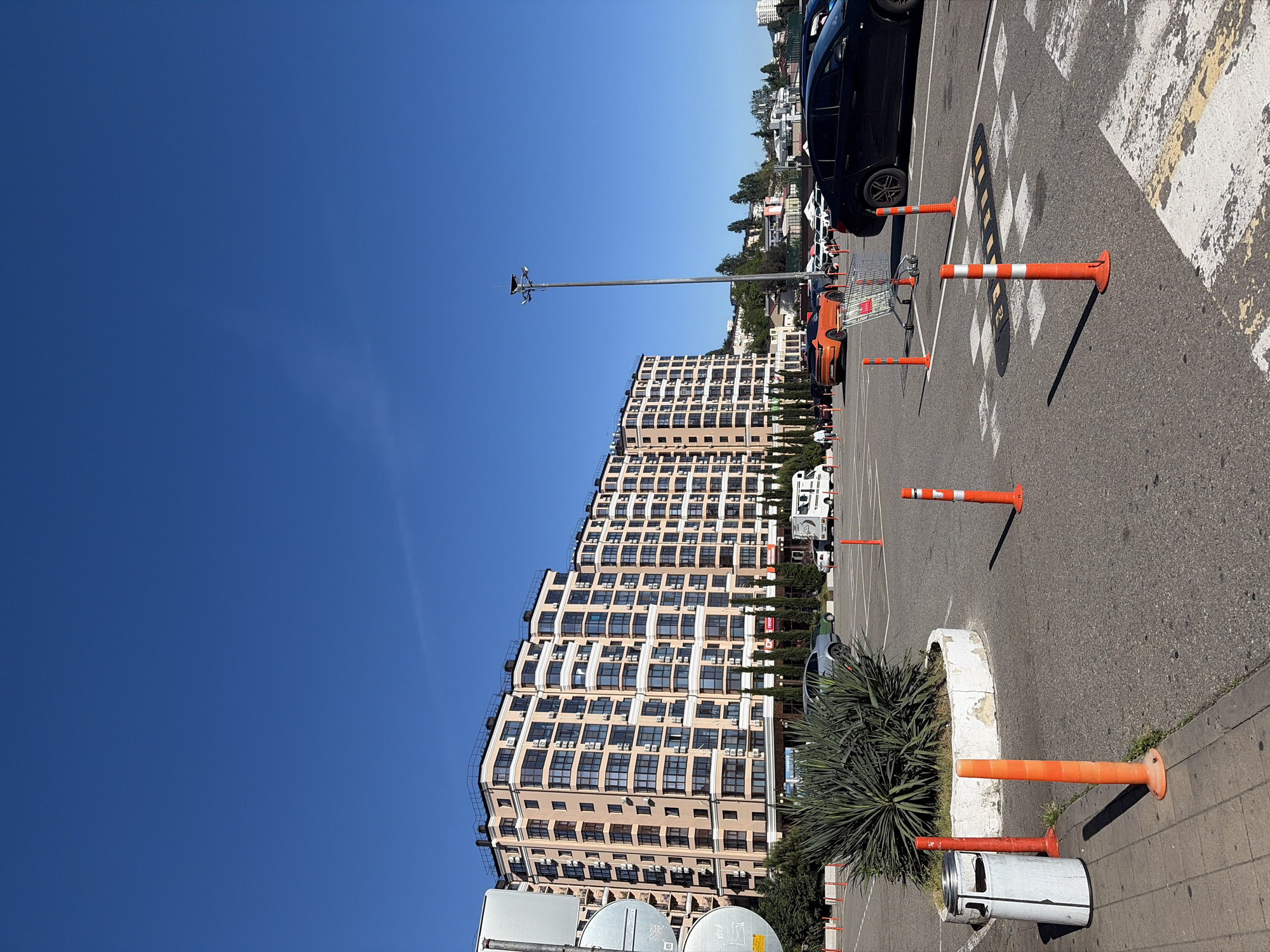 The image depicts an outdoor parking area in Sochi, Russia, under a clear blue sky on a sunny day. The scene is presented from a perspective looking across the parking lot towards a large building and the surrounding environment.
A prominent feature is a multi-story residential or commercial building, characterized by a light beige or peach facade with numerous windows, many of which have air conditioning units mounted below them. The building exhibits a distinctive undulating or wave-like architectural design. In front of the building, there are slender coniferous trees and some palm-like foliage lining the property.
The foreground and right side of the image are occupied by an asphalt parking lot. It features white painted parking space lines, a yellow boundary line, and a speed bump with black and yellow checkerboard patterns. Scattered across the parking lot are several orange traffic cones or bollards, some standing upright and others lying horizontally. Partially visible vehicles include a black SUV, an orange sports car, and a white van, suggesting a mix of parked cars. A shopping cart is also present in the parking area.
In the mid-ground, a tall metal pole equipped with what appears to be security cameras or lights stands out. Towards the bottom right, a white concrete planter or barrier contains a spiky, green, palm-like plant, and a white trash can is situated beside a paved sidewalk. Shadows are sharp and well-defined, indicating strong sunlight, likely in the mid-morning or afternoon. No people are visible in the scene.
The background shows more buildings and dense green trees, hinting at the hilly landscape characteristic of Sochi. Visible text is limited to small, unreadable signs on the building's facade and the markings on the parking lot surface.