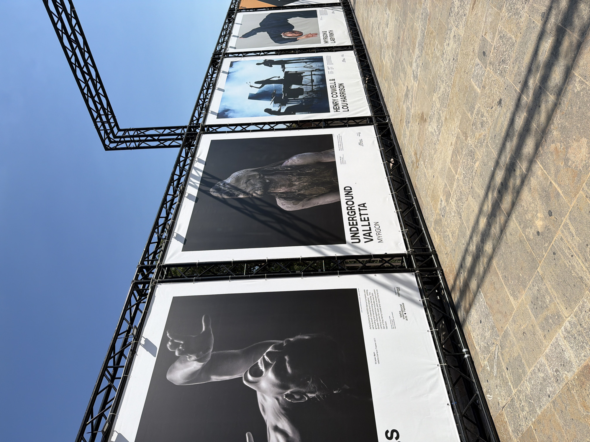 This media file depicts a series of large posters displayed outdoors, likely as part of an arts festival. The posters are mounted on a black metal truss structure against a clear blue sky. The perspective is tilted, looking upwards at the posters and the sky, with a stone-paved ground visible to the right, casting long shadows.
The posters feature artistic imagery and text. The top visible poster, though partially obscured, shows a black and white image of a person in motion. Below it, another poster displays a silhouette of figures on a stage with theatrical lighting, accompanied by text identifying "HENRY COWELL & LOU HARRISON". Further down, a striking black and white image shows a person's head and shoulders with expressive lighting, with the text "UNDERGROUND VALLETTA MYRGON" prominently displayed. The bottom poster is a close-up of a person's torso and arm, captured in a dynamic pose with dramatic chiaroscuro lighting. Text on this poster includes "Malta International Arts Festival" and "FESTIVALS", suggesting the event's name and location. Specific details mention "13 June 8pm Valletta Campus Theatre (VCT)" and describe the performance as "Ashes2Ashes by MOVEO Dance Company".
The scene appears to be set during the daytime, under bright sunlight, indicated by the clear sky and strong shadows. The overall impression is one of an outdoor cultural event, possibly a dance or performance festival, showcasing contemporary art and featuring prominent artists and productions.