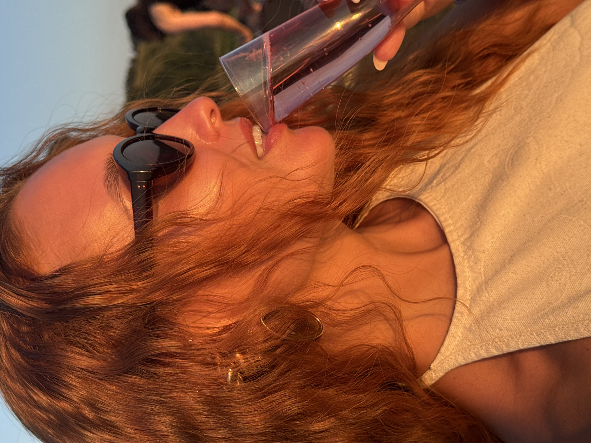 A close-up profile shot shows a person with auburn hair and sunglasses drinking from a glass of pink liquid. The person is wearing a light-colored halter top. The setting appears to be outdoors during sunset, with warm light illuminating the scene. In the background, a blurred figure of another person can be seen. The sky is clear and blue. The image does not provide any specific details about the location in the city of Ольховка, Russia.