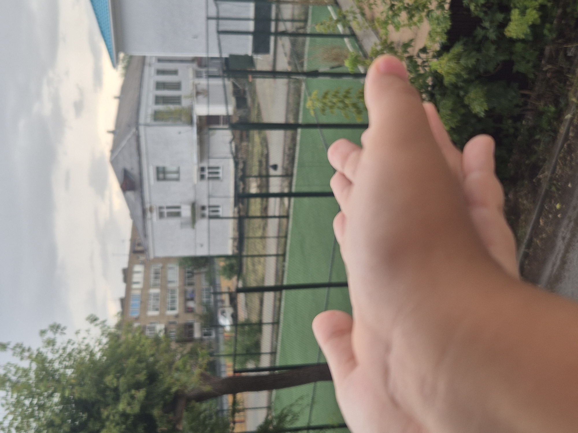 The image shows a close-up of a person's hands in the foreground, with a blurred background of an outdoor sports court and urban buildings. The sports court appears to be a green artificial turf surface enclosed by a dark fence. Beyond the fence, several buildings are visible, including a multi-story residential building with numerous windows, a light-colored two-story building with a pitched roof, and another light-colored building with a bright blue roof. A tree with green foliage is present on the left side of the image. The sky is overcast. The scene appears to be in a city environment, likely a residential or educational area. Without further information or specific landmarks, pinpointing the exact location within Kokshetau, Kazakhstan is not possible.