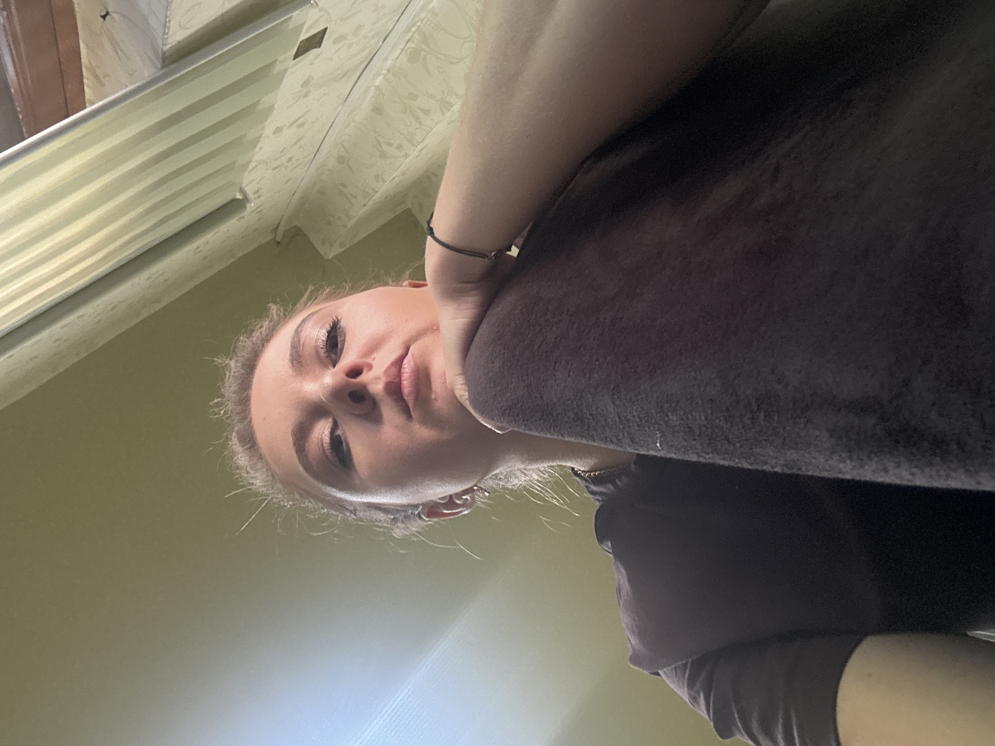 A woman is lying down and looking up at the camera. She is resting her head on her left arm, which is covered by a dark towel. She is wearing a dark t-shirt. The background appears to be a room with a light-colored ceiling and walls. The image is taken from a low angle, looking up at the woman.