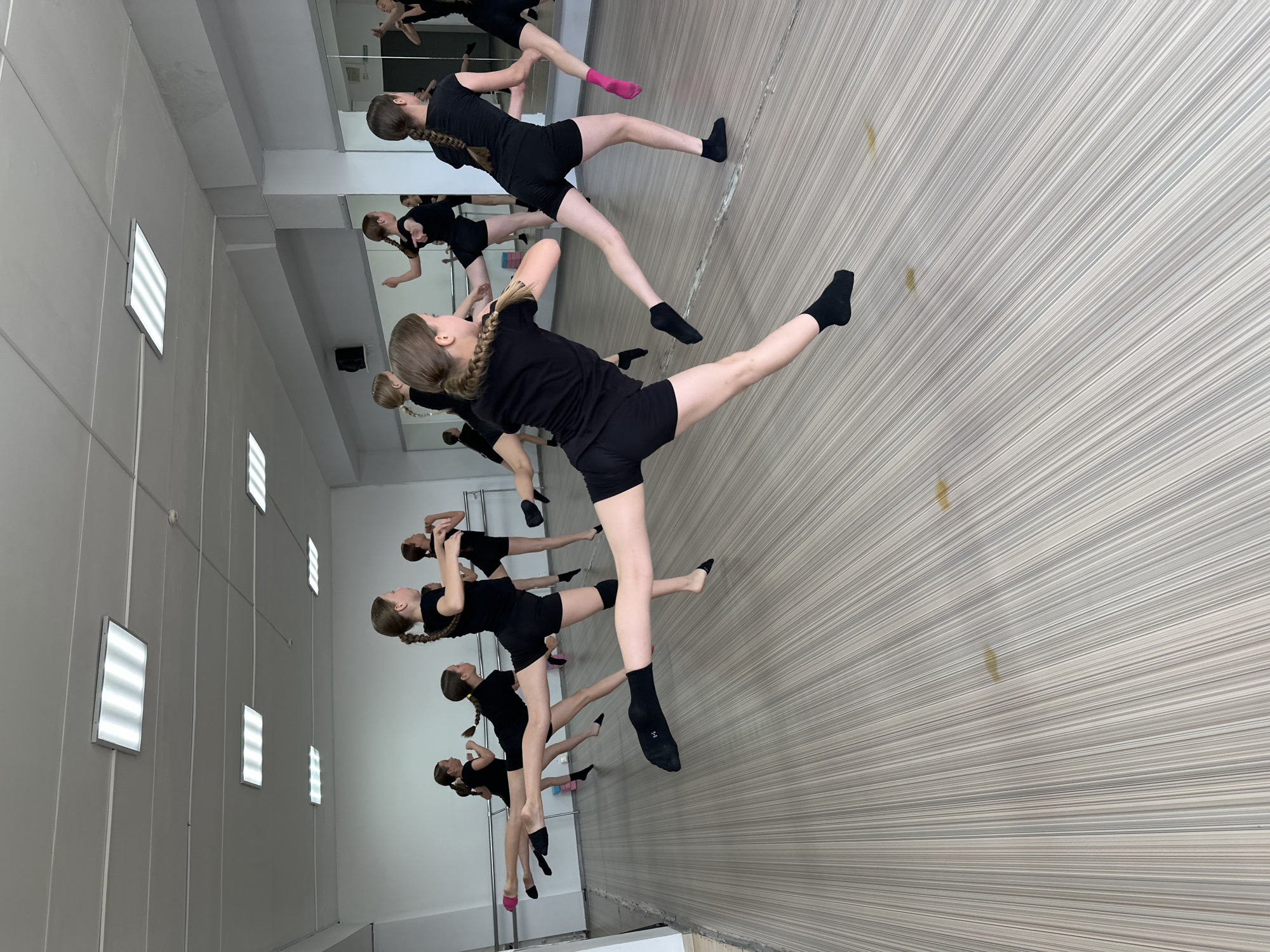The image depicts a dance studio in Votkinsk, Russia, with a group of young girls in black leotards and shorts practicing a dance routine. The girls are captured mid-air, with some executing high kicks and leaps. The studio features a light-colored wooden floor with a striped pattern, white walls, and a mirrored wall on the right side of the frame. Fluorescent lighting fixtures are visible on the ceiling. A ballet barre runs along the wall. The overall atmosphere suggests a professional dance class, focusing on athleticism and coordinated movement.