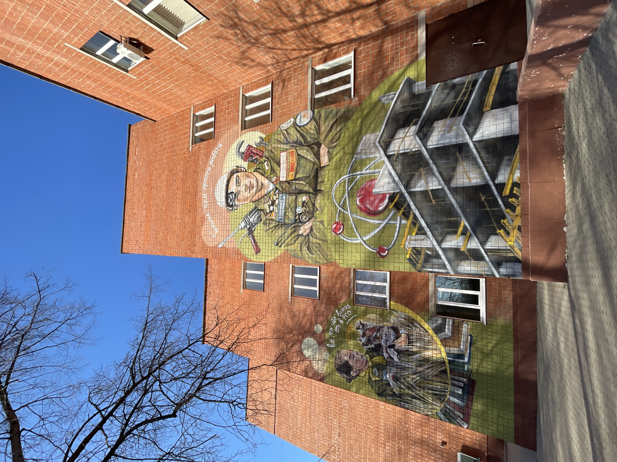 The image shows a brick building in Izhevsk, Russia, adorned with a large mural. The mural depicts two men in military attire, interspersed with images of scientific equipment like an atom model and a metal framework. The mural seems to be a tribute to the military and scientific community. The building has rectangular windows arranged at different levels. In the foreground, there are bare tree branches against a clear blue sky. A portion of the street with shadows is visible on the right side.