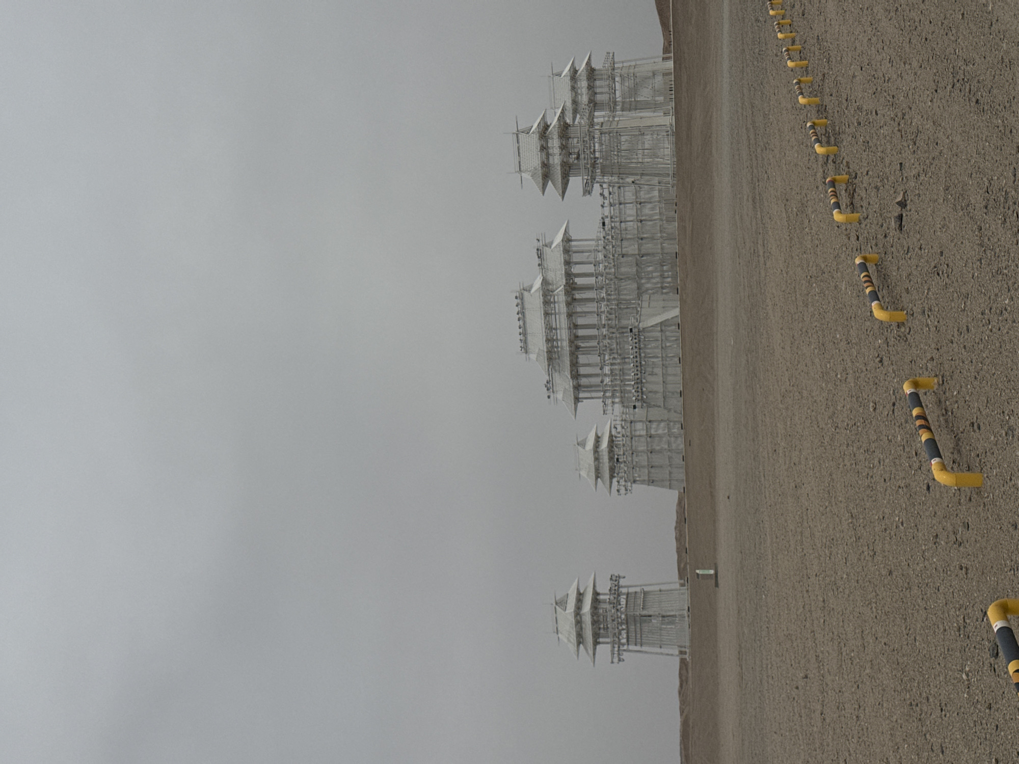 The image depicts a desolate landscape under a gray sky. Situated in Guazhou County, China, the scene features a row of structures reminiscent of pagodas, constructed from white scaffolding and platforms. The ground is arid and beige, marked by evenly spaced, yellow and black parking bollards. The buildings appear incomplete and unused. No people or activities are visible.