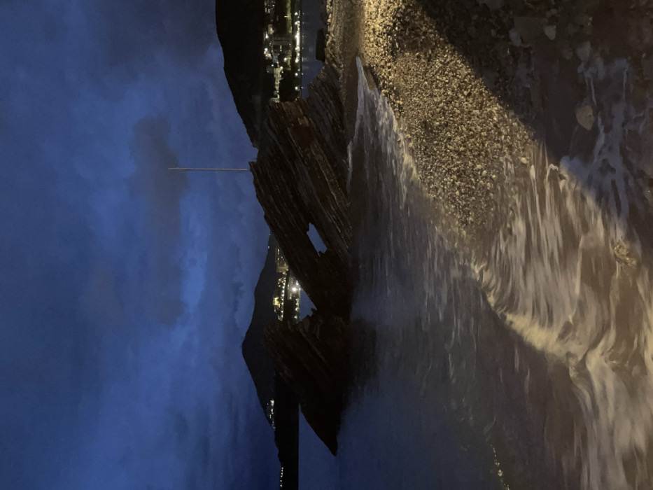 Night scene of a beach with dark blue sky and clouds. A tall rock formation stands on the left, an archway visible within its structure. Lights from a town are seen in the distance. The shoreline has small pebbles. Waves roll onto the shore.