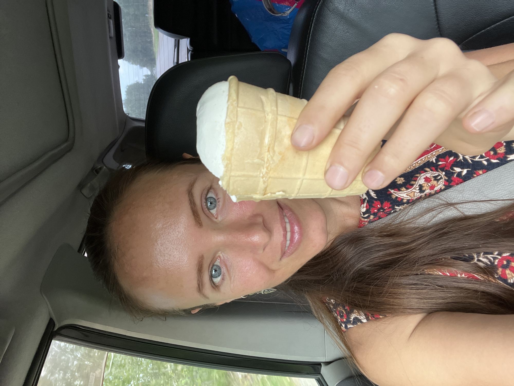 A woman with blue eyes and brown hair is sitting in the passenger seat of a car, holding an ice cream cone. She is wearing a dress with a red and black floral pattern. The car appears to be on a road in a rural area, with trees visible outside the windows. The weather is overcast. The location is identified as Miasskoye, Russia.
