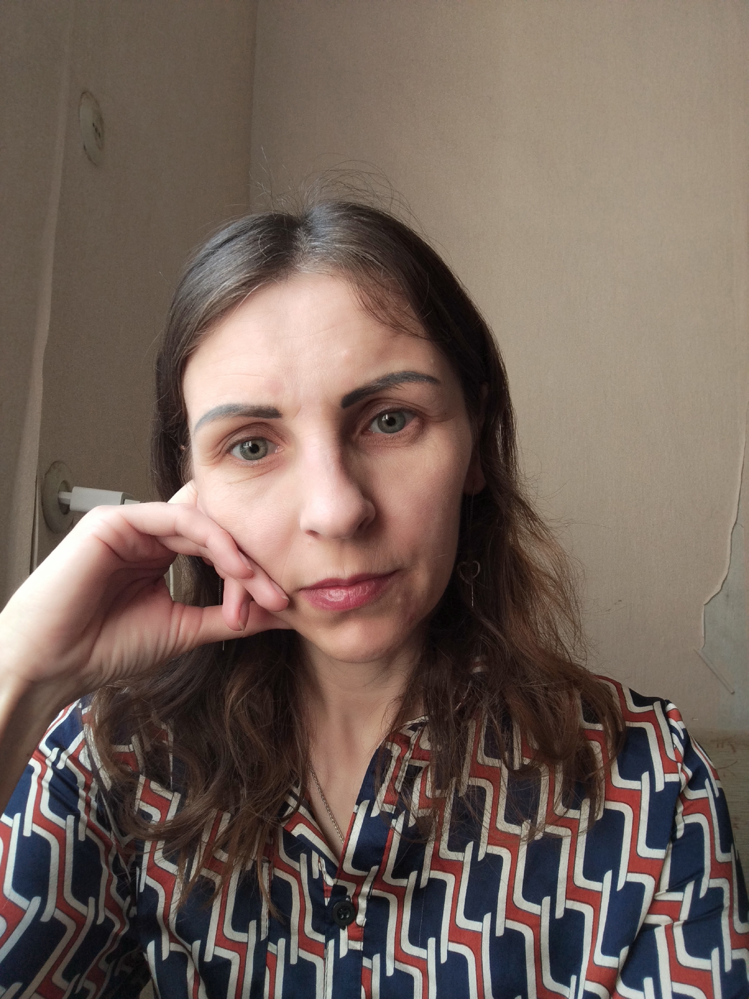 A close-up, waist-up, indoor portrait photograph shows a woman with medium-length, brown, wavy hair. She is looking directly at the camera, with her left hand resting on her cheek and chin, supporting her head. Her eyes are green, and she has dark eyebrows. She is wearing a dark blue, patterned shirt with geometric designs in red and white. A thin silver chain necklace is visible. The background appears to be a neutral-colored wall with some texture and a visible light fixture on the left side. The image is likely taken in Vinnytsia, Ukraine.