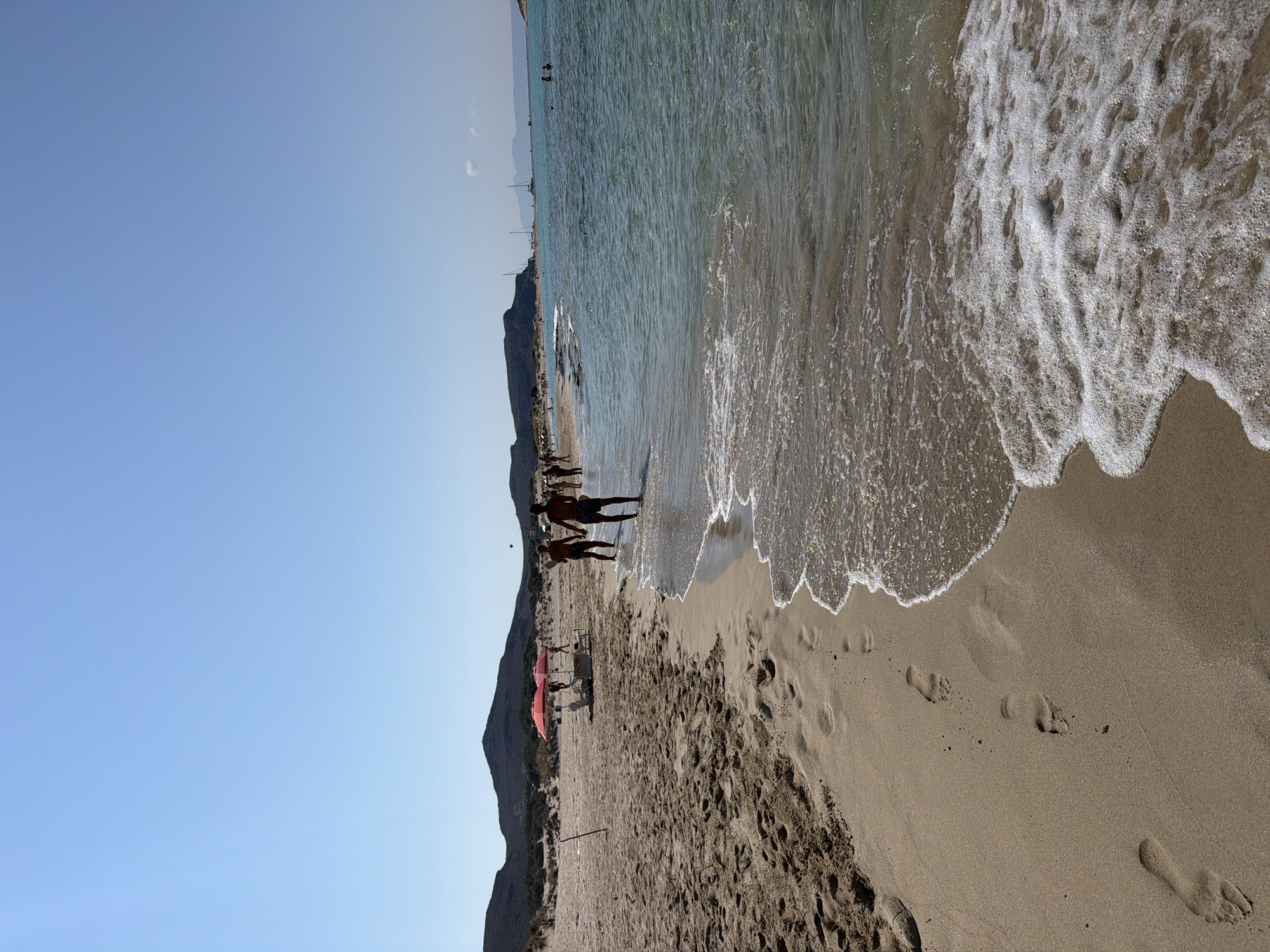 This image depicts a sunny day at a beach in an unidentified location in Greece. The foreground is dominated by a sandy beach with numerous footprints leading towards the water. The ocean is a clear, turquoise color with gentle waves lapping at the shore. In the middle ground, several people are visible enjoying the beach. Two individuals are standing in the shallow water, appearing to play a game. Further down the beach, there are red umbrellas providing shade for beachgoers, along with lounge chairs and a table. In the background, a chain of low, rocky hills forms the coastline, and several sailboats are visible on the horizon. The sky is a bright, cloudless blue. The overall scene conveys a sense of leisure and relaxation on a warm summer day.