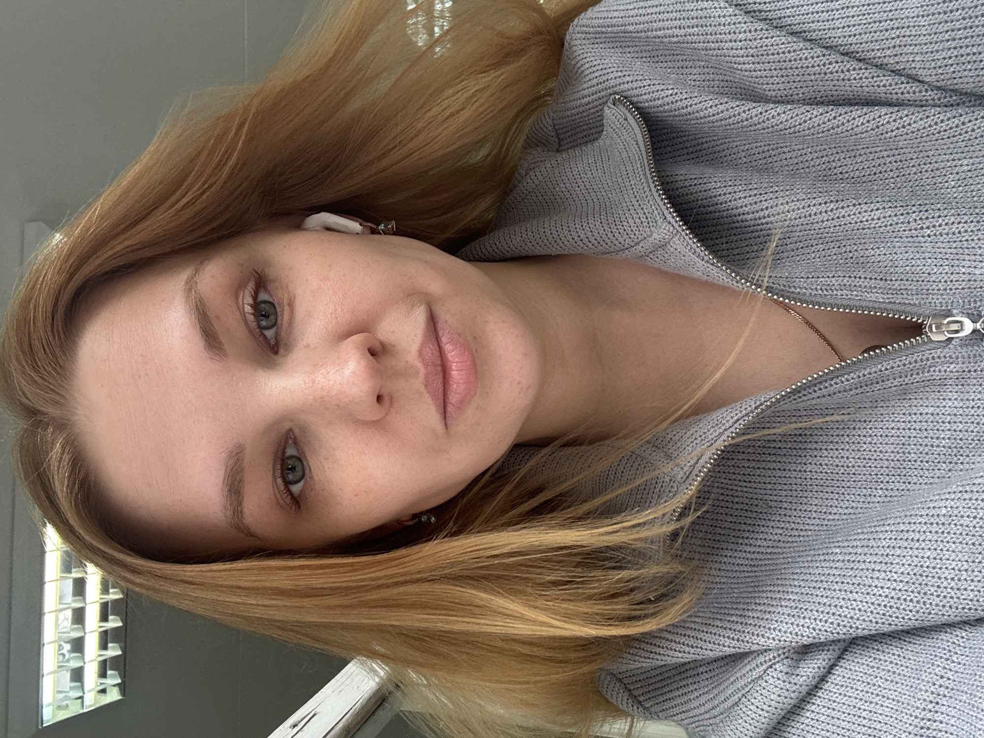 A close-up photograph shows a fair-skinned woman with blonde hair wearing a gray zip-up sweater. An earbud is visible in her left ear. A window with a multi-pane grid is visible in the background. The location is not discernible from the image. Rusanovo, Russia is not identifiable in the image.