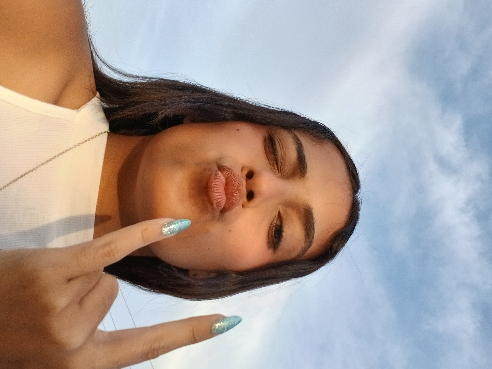 A woman is taking a selfie with the sky as the background. She is wearing a white tank top and a silver necklace. She is making a kissing face and holding up her index and middle fingers. Her nails are painted light blue and sparkle. The sky is light blue with white clouds. The location cannot be determined.