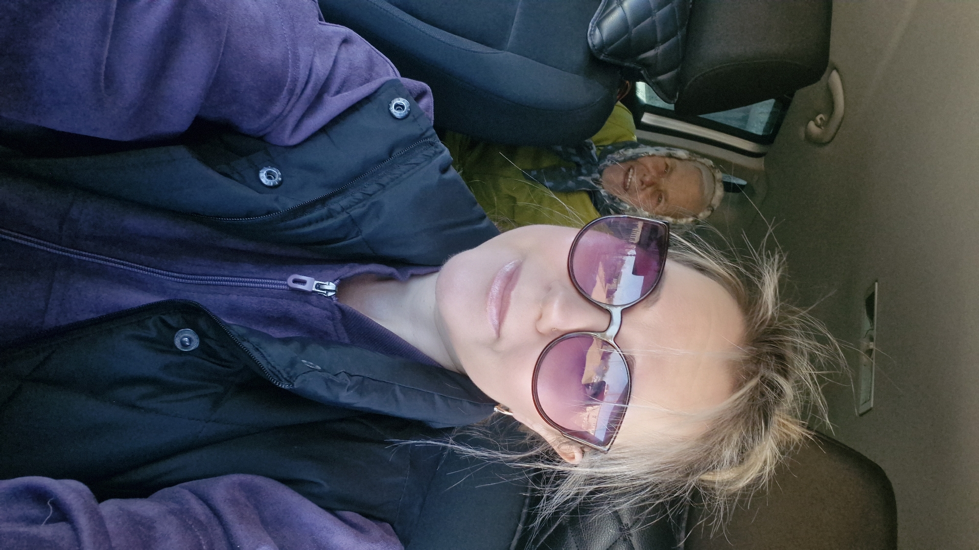 An indoor, vertical selfie captures two individuals inside a car. In the foreground, a person with blonde hair and wearing large, pink-tinted sunglasses is seen from the chest up. They are wearing a purple hooded garment underneath a black puffer vest, which is partially unzipped. Their lips are slightly parted, and their expression is neutral.
Behind and to the right of the foreground subject, partially obscured by the car seat and reflected in the sunglasses, another person is visible. This individual has light-colored hair and is wearing a yellow or lime-green jacket with a fluffy or fur-trimmed hood. They are smiling broadly, looking directly towards the camera.
The setting is the interior of a car, indicated by the black car seats, a quilted headrest, and part of the car's ceiling and window in the background. Light from outside suggests it is daytime. The heavy clothing worn by both individuals implies cooler weather. There is no visible text in the image. The location is contextualized as Otchor, Russia.