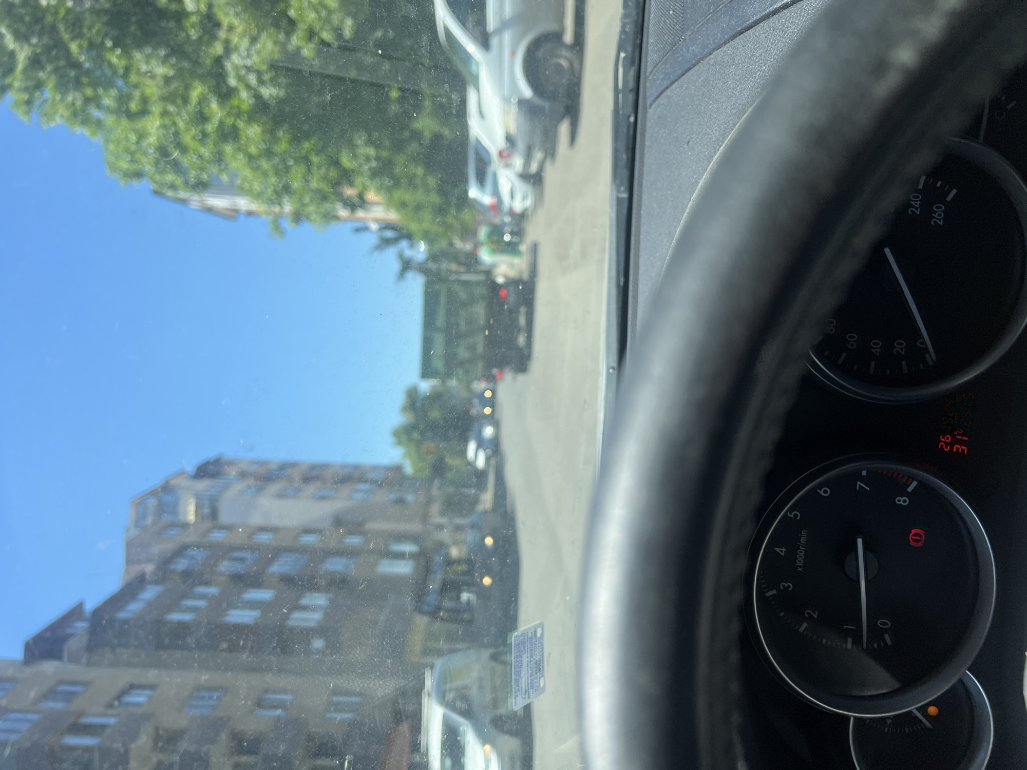 Here's a detailed description of the image:
**Scene Overview:** The image is taken from inside a car, looking out through the windshield. The car appears to be stopped or moving very slowly on a city street. The dashboard and steering wheel of the car are prominent in the foreground.
**Environment:**
* **Setting:** Urban, likely a residential or commercial area.
* **Weather:** Sunny, clear sky.
* **Road:** Appears to be a paved street with other vehicles parked along the side.
**Objects:**
* **Car Interior:** Steering wheel, dashboard with speedometer and tachometer (showing the car is idling), temperature display.
* **Exterior:** Buildings (likely apartment blocks), parked cars, trees.
* **License Plate:** Partially visible license plate in the center view out the windshield.
**Location (Inferred from OCR and Visual Cues):**
While the image doesn't explicitly state the location, the details of the image combined with the additional information from the OCR lead to the strong suspicion that it is located in **Burgas, Bulgaria**. I can identify no specific landmarks to confirm the location more precisely.