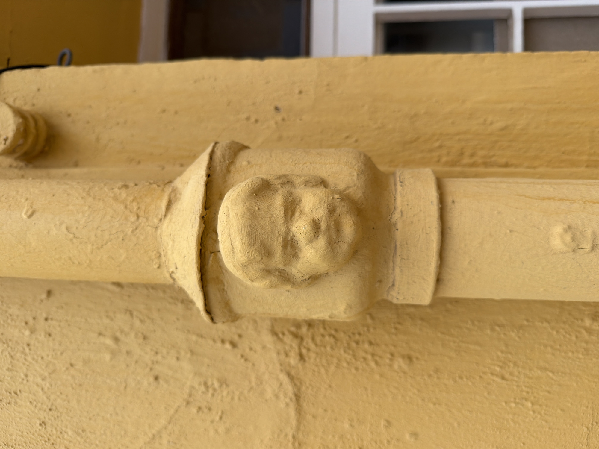 A close-up, eye-level shot focuses on a decorative architectural element, specifically a yellow, textured downspout with a sculpted cherub face embedded within its structure. The downspout is attached to a building with a similar yellow, textured facade, and a portion of a white-framed window can be seen in the blurred background to the right. The cherub face is rendered in a plaster-like material and appears to be aged, with some visible surface imperfections and textural variations. The overall impression is one of an old, possibly historical, building in Castelló De La Plana, Spain, where such decorative details are often found.