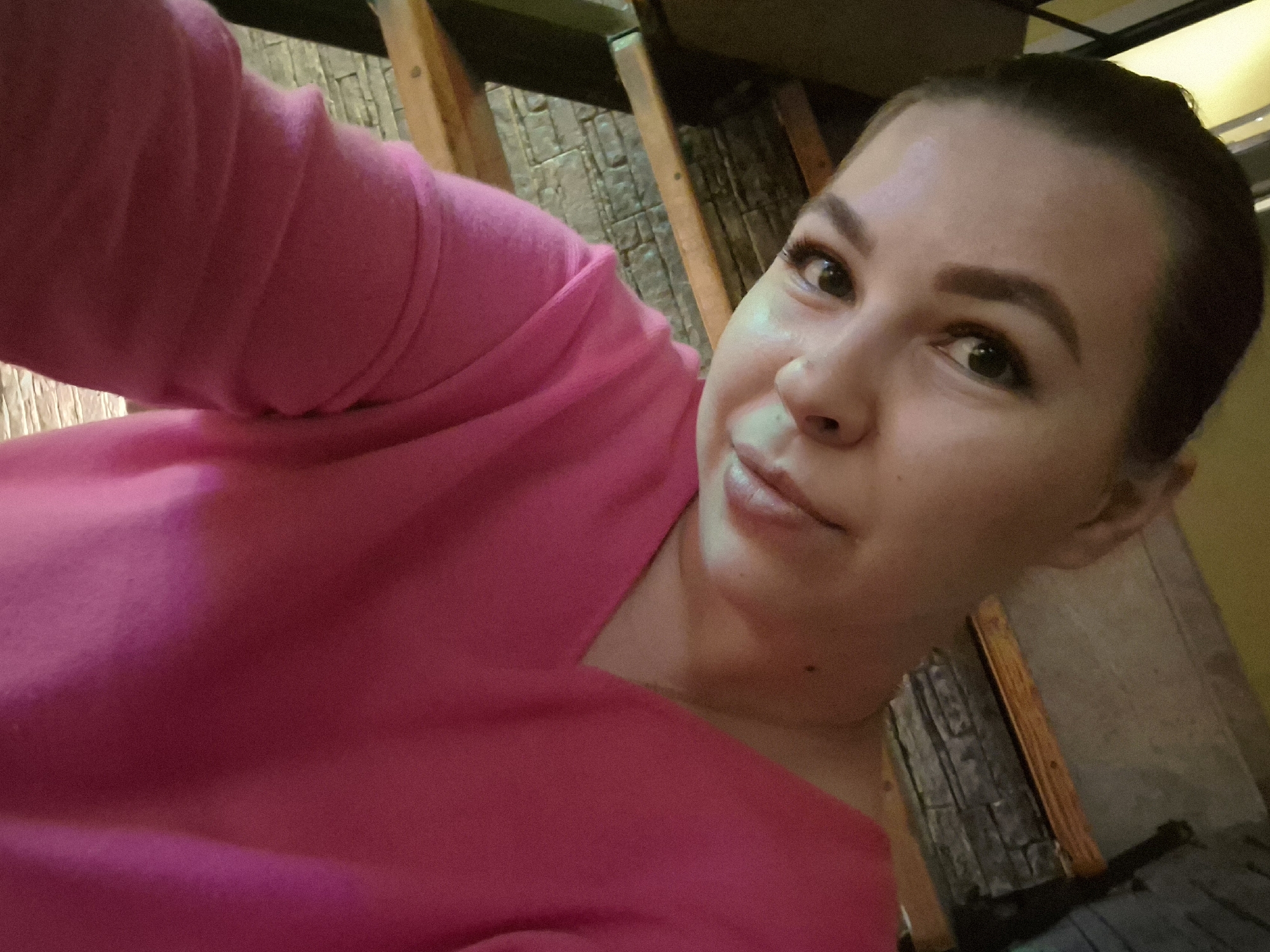 A woman with fair skin and dark hair pulled back is posing for a selfie. She is wearing a pink top. The background appears to be an interior space with stone-like paneling on the walls. Some wooden beams or supports are visible. The location is indicated to be Chaikovsky, Russia.