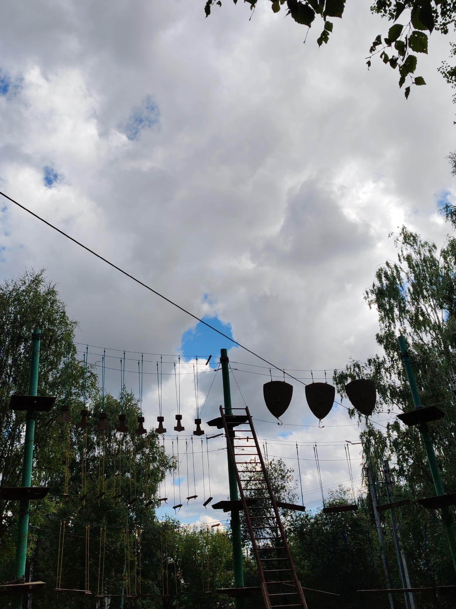 The image shows an outdoor ropes course, likely in a park or recreational area. In the foreground, several green metal poles support various elements of the course, including wooden platforms and ropes with suspended obstacles. A rusty metal ladder extends upwards from the bottom right towards the middle of the frame. Above the course, a series of wires are strung across the scene. Three shield-shaped obstacles hang from the wires in the upper center of the image. Lush green trees surround the ropes course. The sky above is cloudy with patches of blue sky visible. The scene appears to be captured on a bright day with diffused sunlight.
This specific ropes course is located at "Park Zarechny" in Syktyvkar, Russia. The park offers various attractions for visitors, including this adventure park.