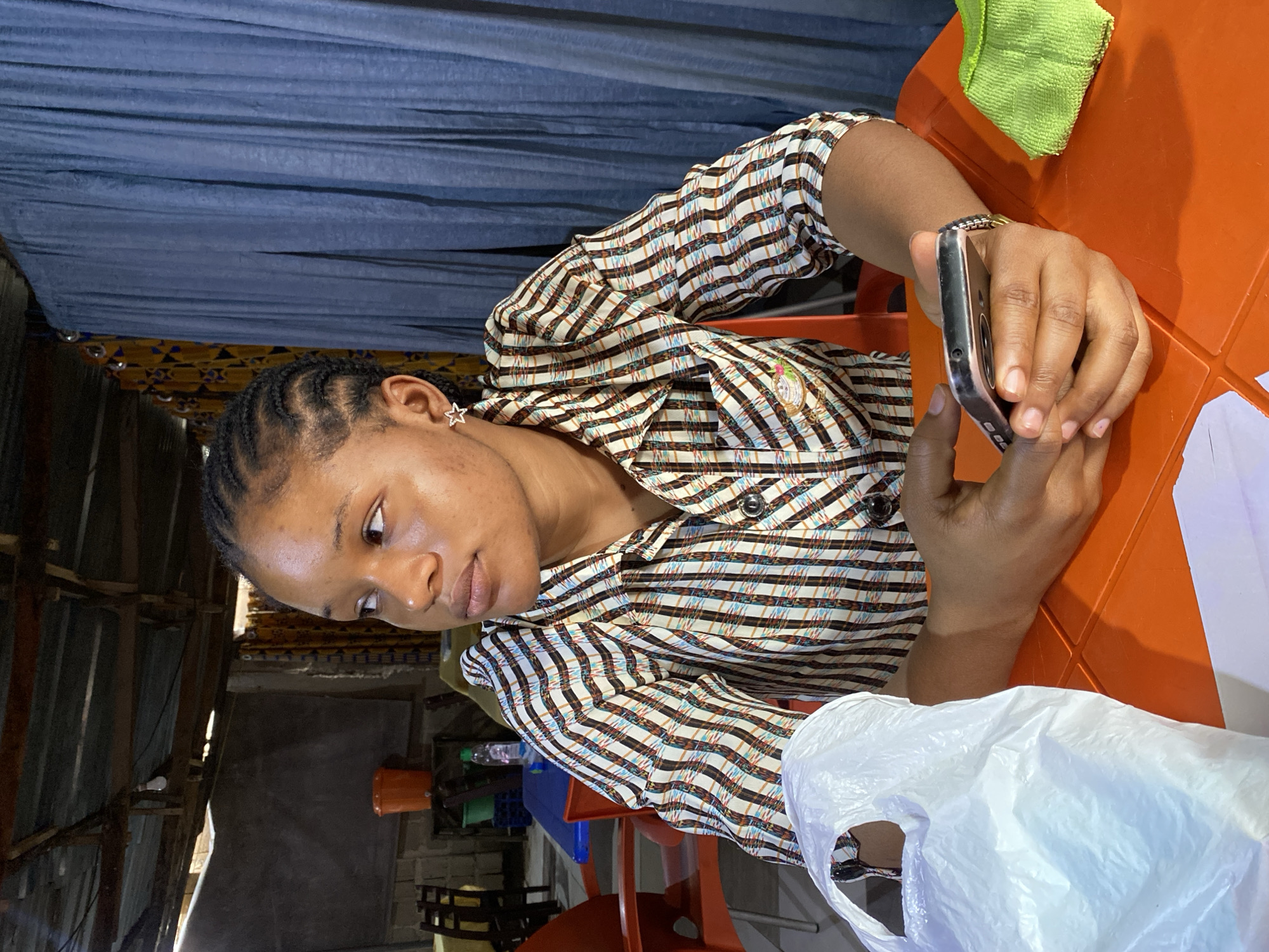 A young woman with braided hair is sitting at an orange table, looking at her smartphone. She is wearing a striped long-sleeved shirt and a star-shaped earring. A green cloth is draped over the corner of the table. In the background, a blue curtain hangs, and beyond that, there is a glimpse of a corrugated metal structure. The scene appears to be indoors, possibly a casual outdoor or semi-enclosed space, given the visible construction elements and the general lighting. The woman's expression is focused on her phone. A white plastic bag is also visible on the table in the foreground. The location context is Jos, Nigeria. There is no visible text in the image.