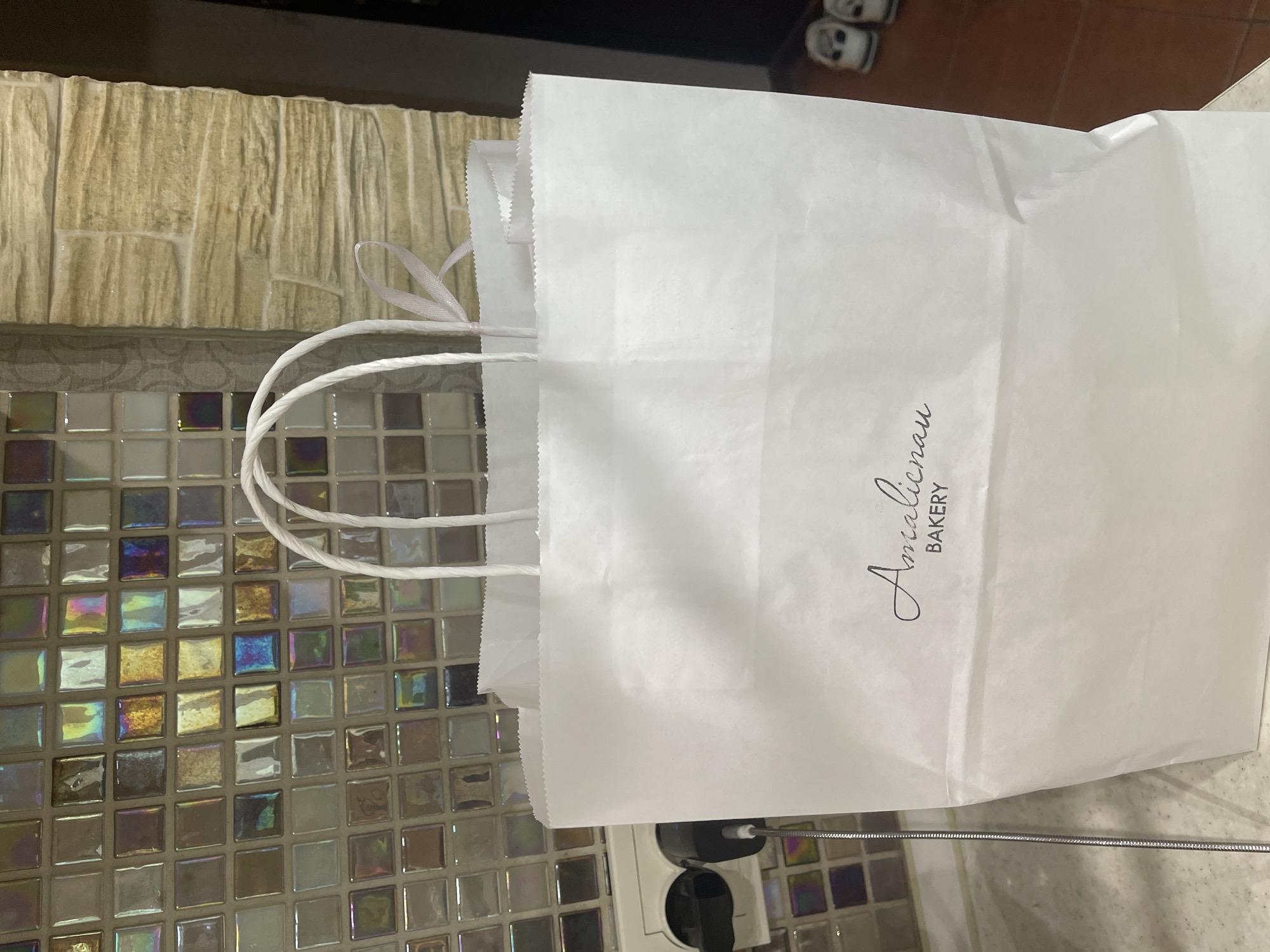 A white paper shopping bag with "Amalienau BAKERY" printed on it in black script stands on a countertop. The bag has white rope handles and a small pink ribbon tied to one of the handles. The background features a wall tiled with small, iridescent square tiles and a textured, stone-like wall. To the left of the bag, a white electrical outlet with a black plug is visible. In the background to the right, two white shoes are on the floor. The image is likely taken indoors, possibly in a kitchen or retail space. The location is identified as Kaliningrad, Russia.