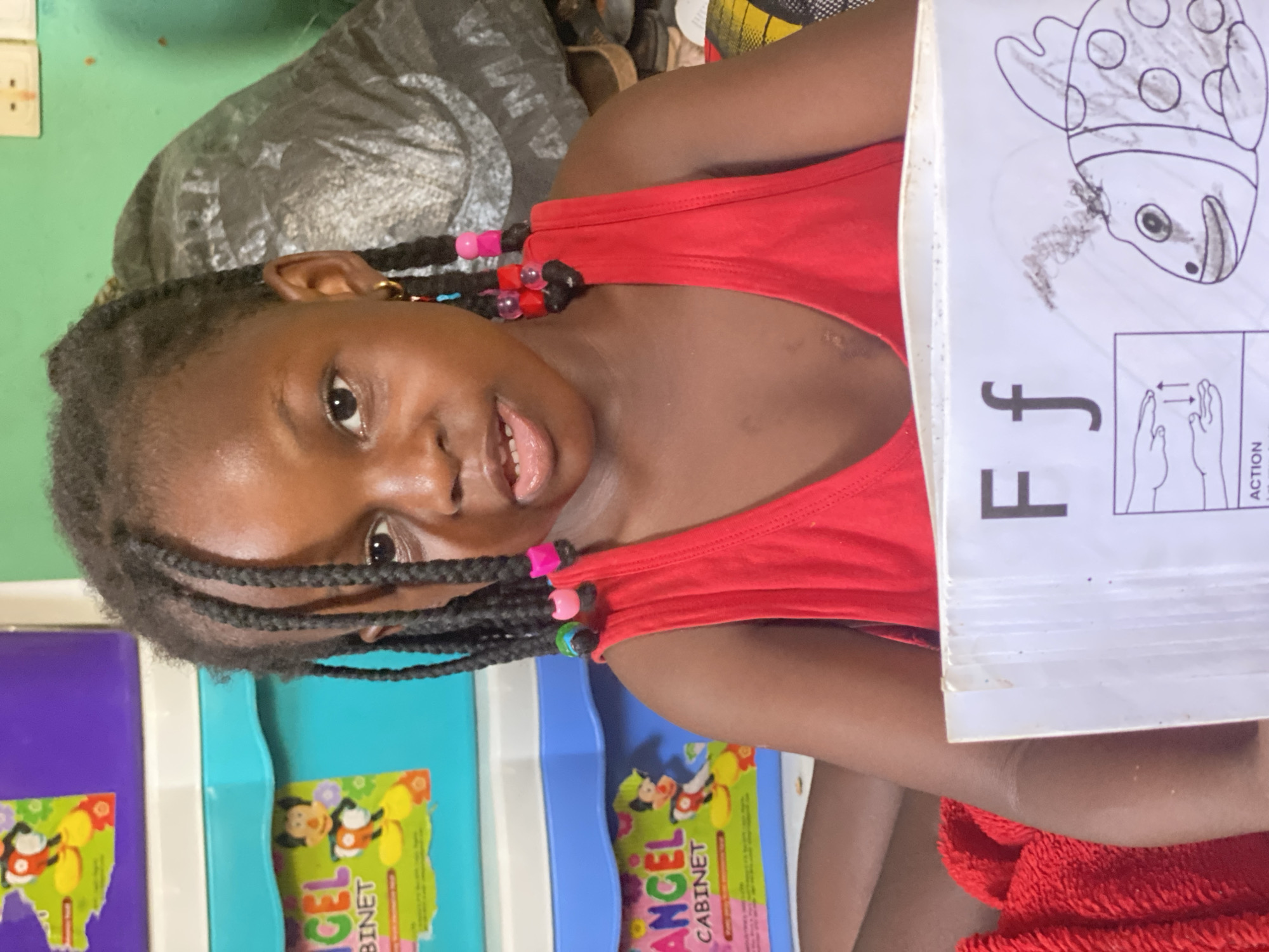 A young Black girl with braided hair adorned with colorful beads is positioned indoors, likely in a home setting. She is wearing a red tank top. The girl is holding a learning book open to the letter "F" with an illustration of a fish. In the background, there are children's storage bins and what appears to be a grey blanket. The location is identified as Rido, Nigeria.