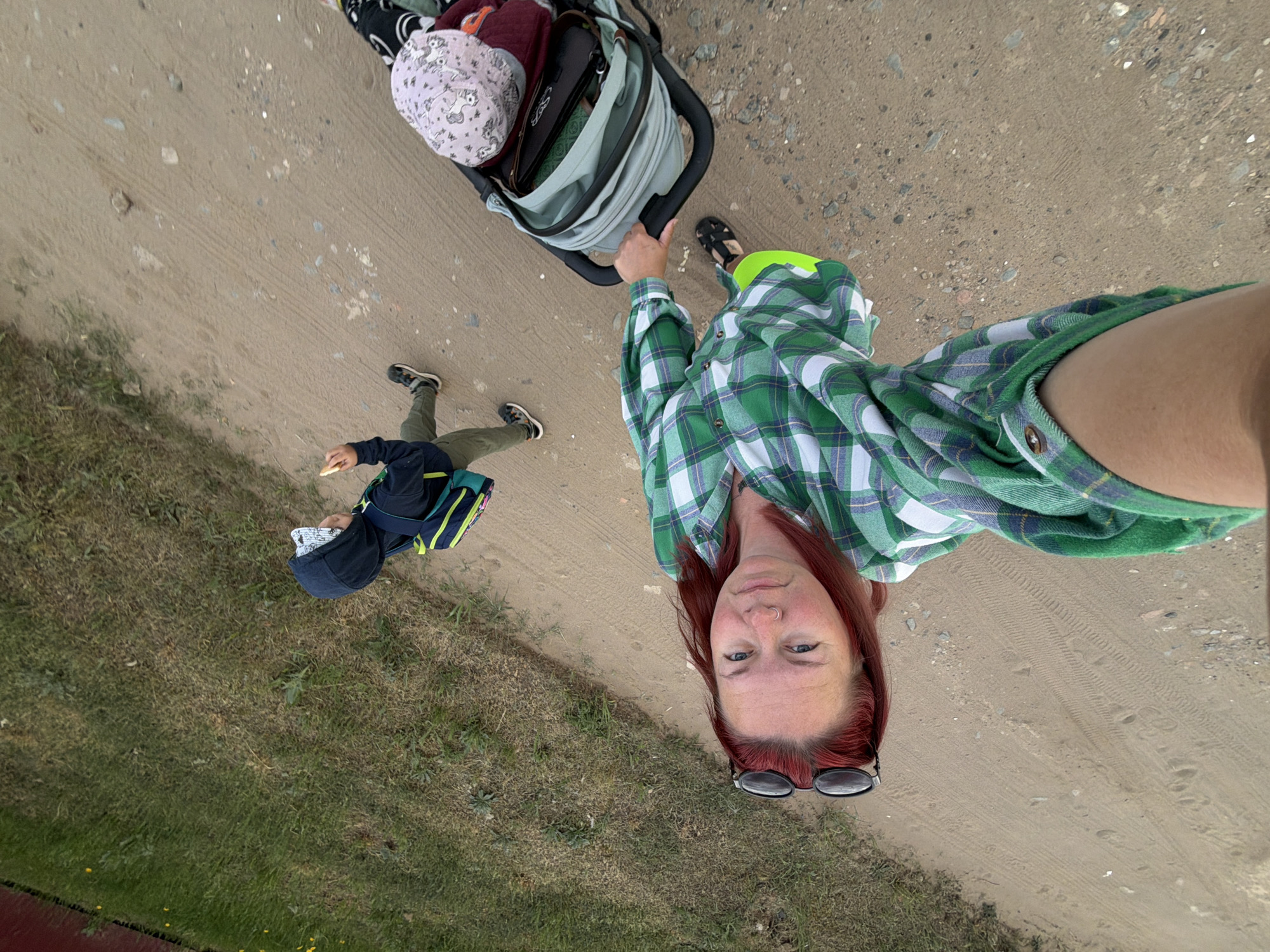 An outdoor eye-level, close-up selfie shows a woman and a child on what appears to be a dirt path in Chaikovsky, Russia. The ground beneath the dirt path has areas of green grass. The woman is wearing a green plaid shirt and black sunglasses. Her hair is a faded red color. She is looking directly at the camera with a neutral expression. A stroller containing various items sits to her left. A child wearing a dark blue hooded jacket and a backpack walks alongside her to her left. The child appears to be eating. Both the woman and the child are wearing black shoes. The background is out of focus, but appears to be a natural environment. The photo is oriented vertically.