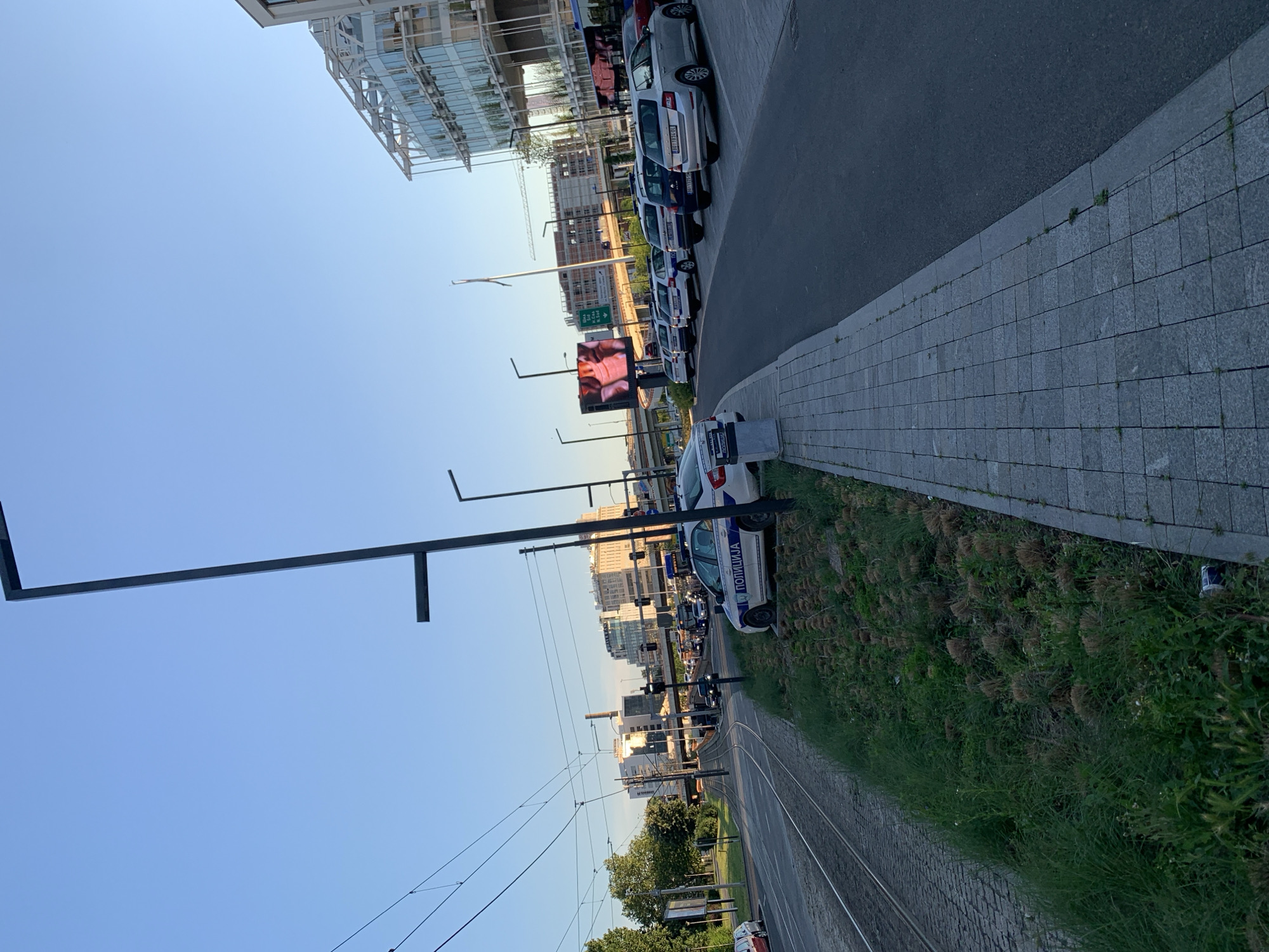 The image shows a street scene in Belgrade, Serbia. A line of police cars is parked along the roadside, beside a grey, cobblestone sidewalk. Between the sidewalk and the tram tracks is a narrow strip of greenery. Overhead tram wires are visible. A large digital billboard displaying a person's face is on the opposite side of the road. Buildings line the street in the background. The sky is clear and blue. The image suggests a street setting, possibly with a focus on security or law enforcement activity.