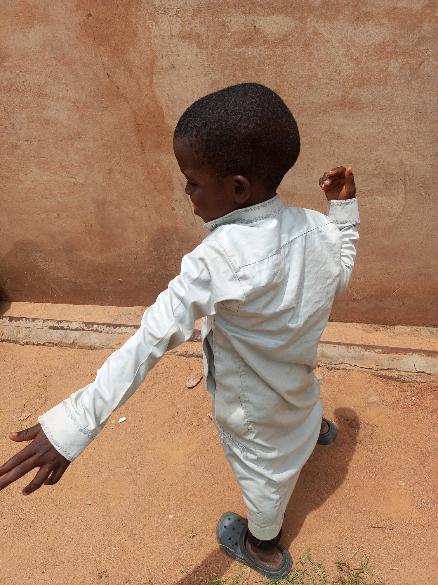A young boy is standing in front of a textured, terracotta-colored wall, possibly in Tempure, Nigeria. He is wearing a pale blue long-sleeved shirt with decorative stitching on the collar and cuffs, and dark trousers. His right arm is extended to his side, with his hand open, while his left arm is bent at the elbow, with his fist raised. The ground is covered in reddish-brown sand with scattered blades of grass. He is wearing dark, slip-on shoes. The lighting suggests it is daytime, with a noticeable shadow cast by the boy. The wall has several visible cracks and imperfections.