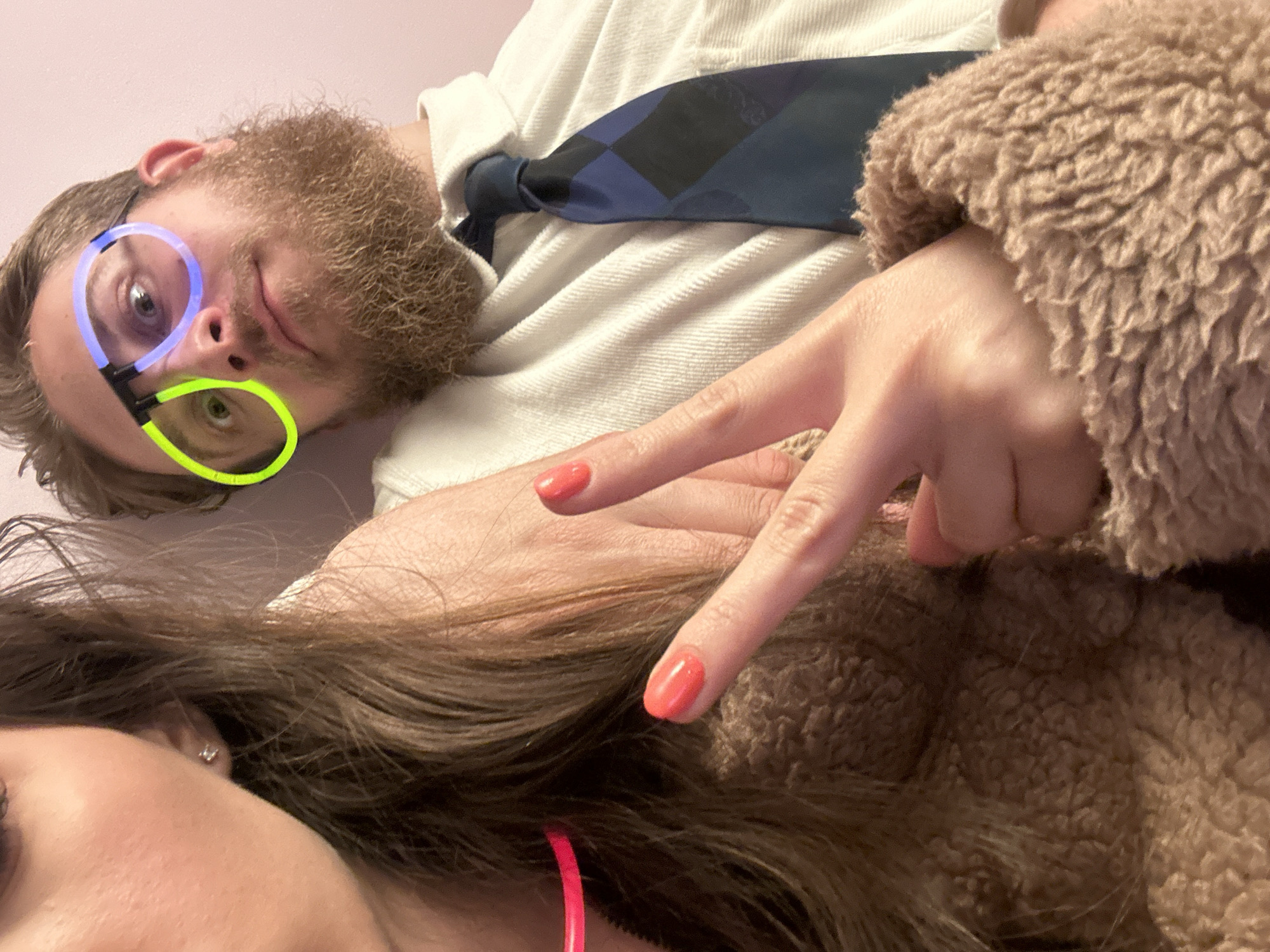 A selfie captured indoors features a man and woman. The man, positioned in the upper portion of the frame, wears glowstick glasses with blue and green rims. He has a beard and a tie. The woman occupies the lower portion of the frame. A hand with orange nail polish makes a peace sign. A pink necklace encircles the woman's neck. The setting appears to be indoors, with a light pink wall. The location of Konska, Bulgaria is not discernible from the image.